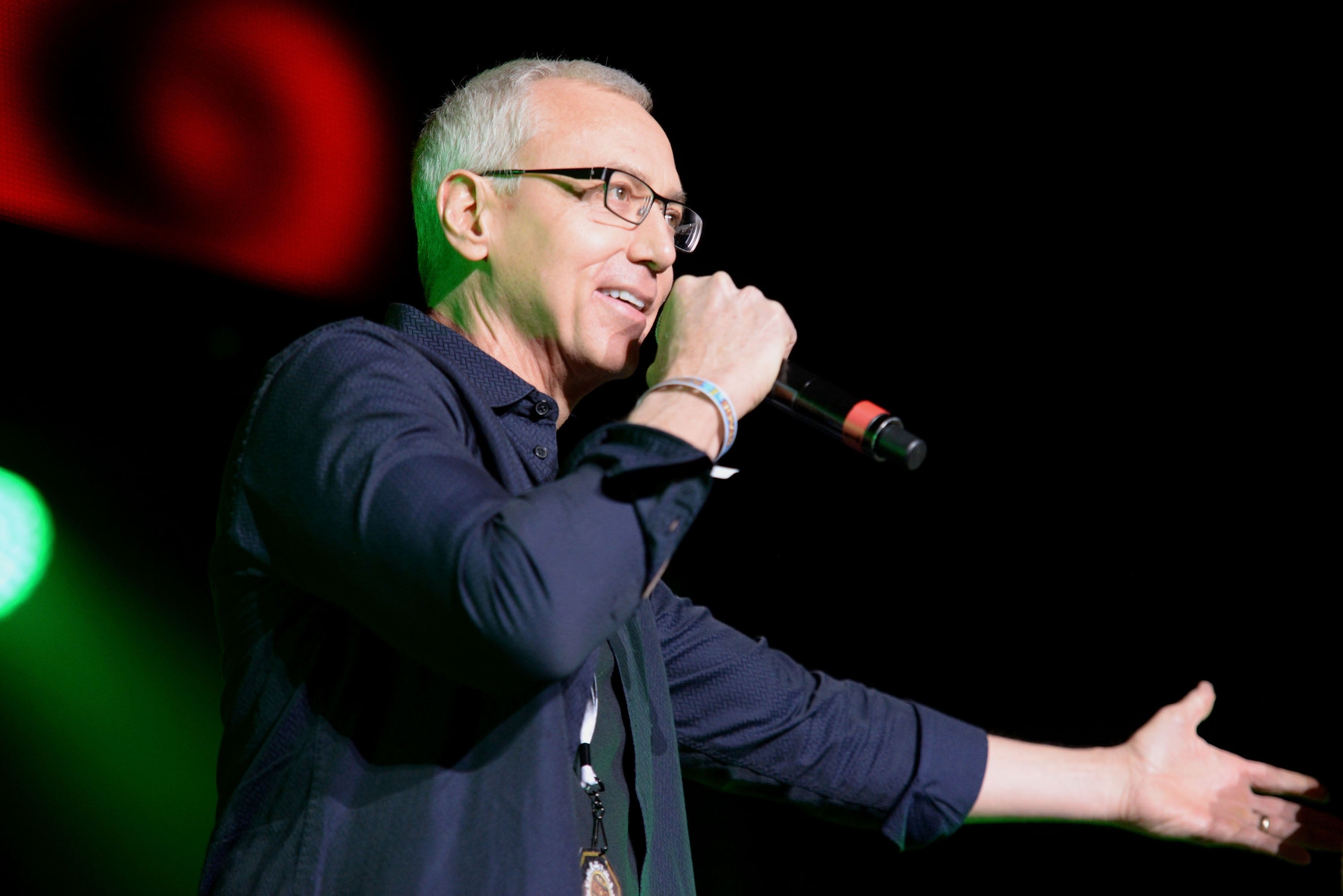 Dr. Drew Pinksy with glasses speaking into a microphone connected  stage, gesturing with 1  hand, dressed successful  a casual shirt