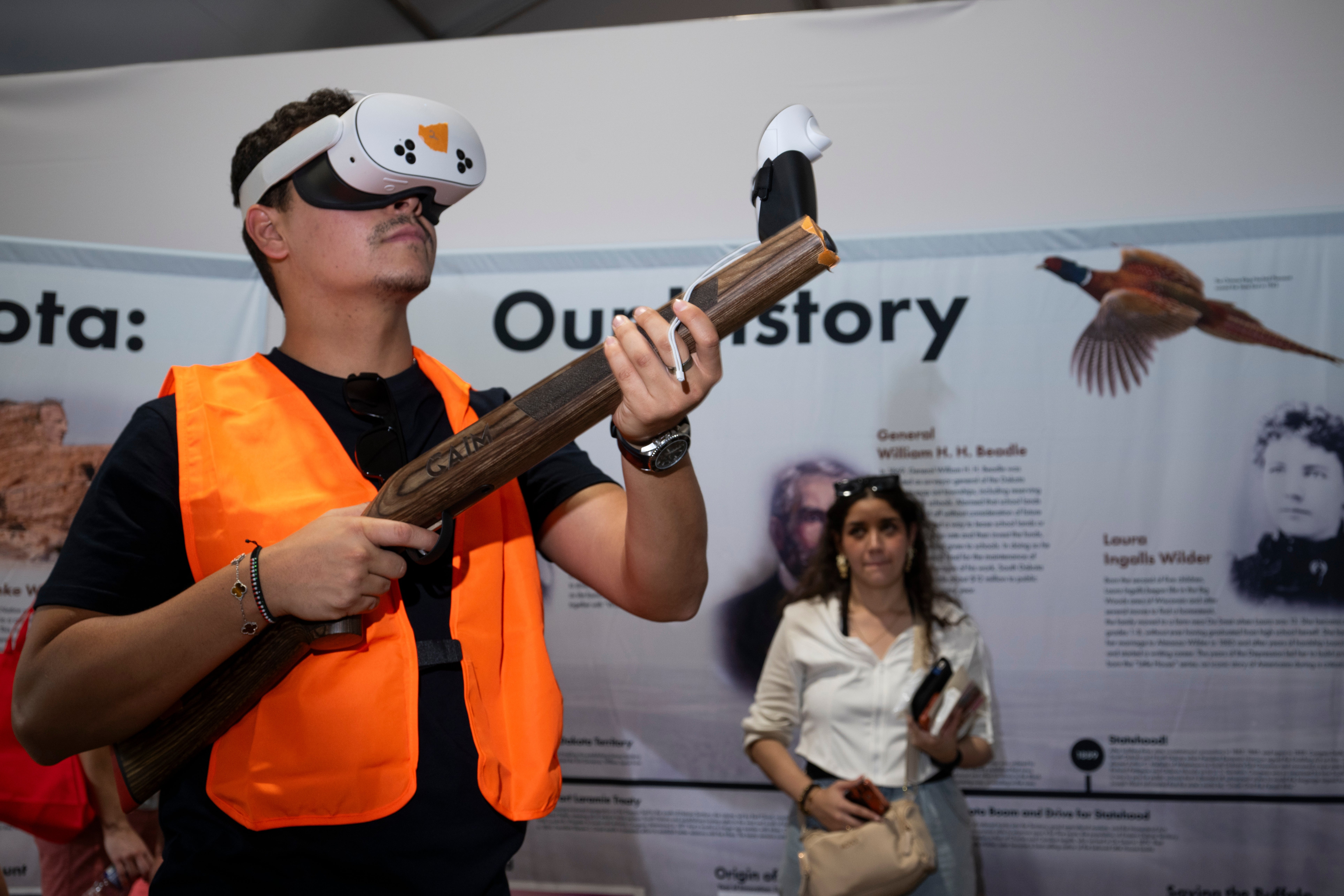 Person som har på seg VR-headset og oransje vest, holder en falsk trerifle, simulerer et jaktspill. Andre deltakere og en skjerm er i bakgrunnen