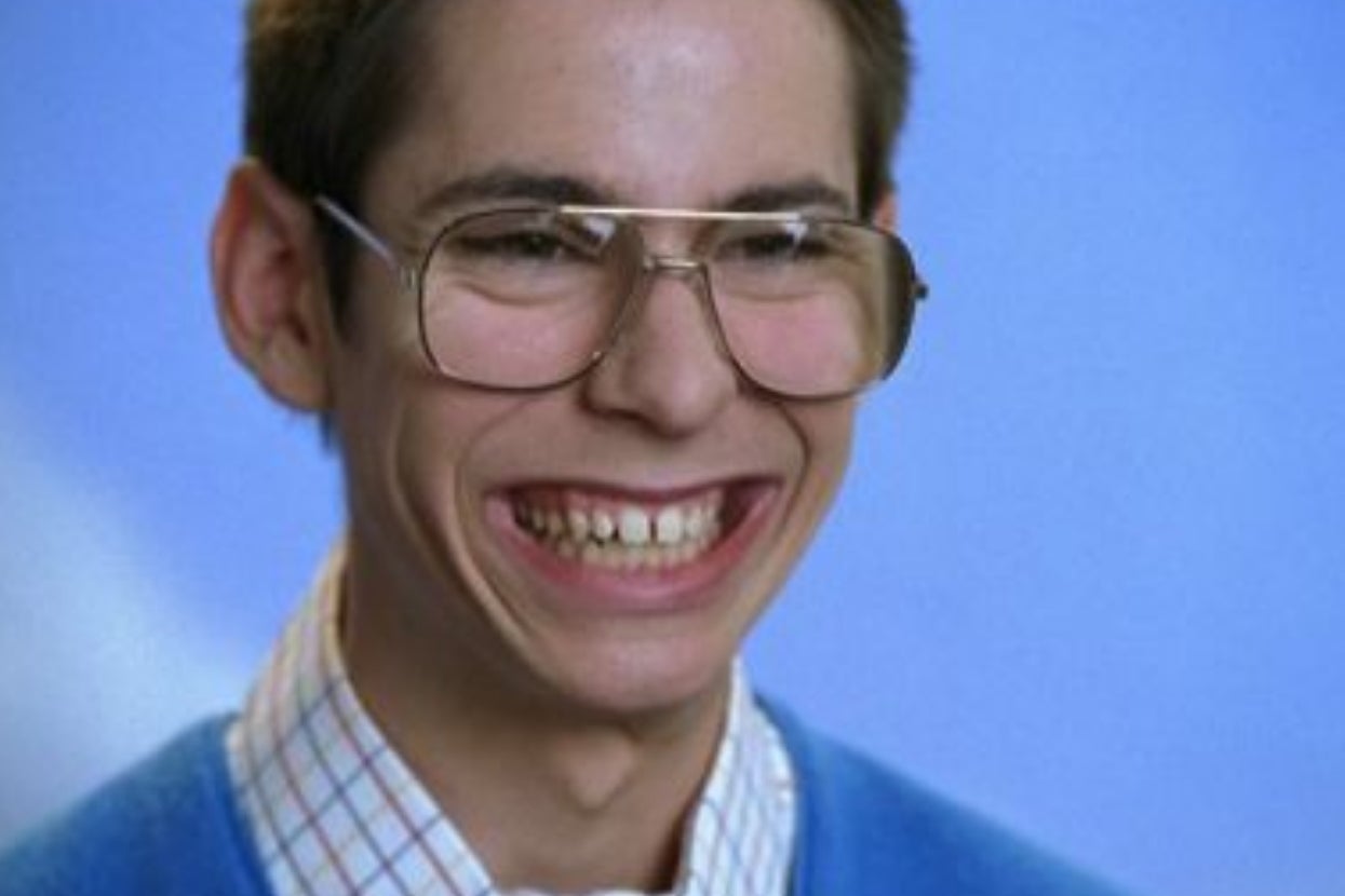 Person smiling broadly, wearing large glasses, a plaid shirt, and a sweater, set against a plain backdrop