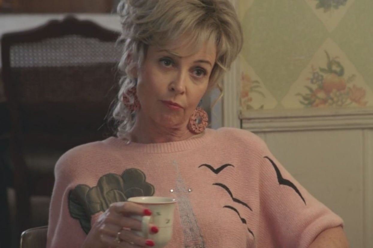 Person with a retro hairstyle holds a teacup, wearing a sweater with a bird and Eiffel Tower design, sitting indoors with a thoughtful expression