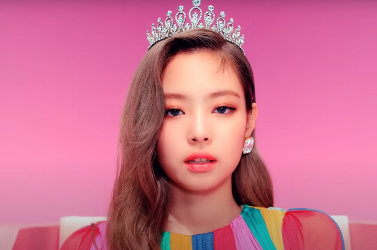Jennie from "BLACKPINK" in a music video wearing a sparkling tiara and vibrant, multicolored attire, set against a plain backdrop