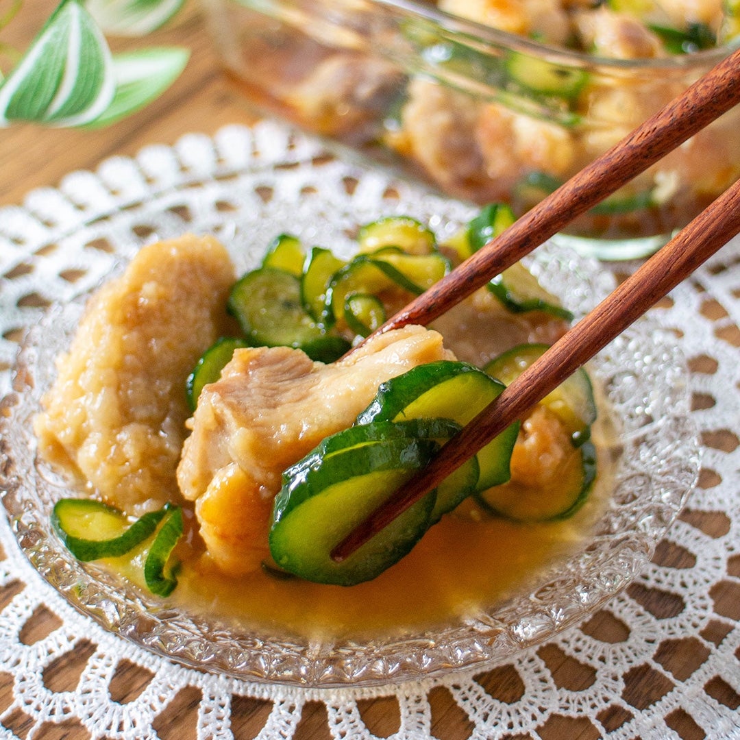 ガラス皿に盛られた鶏肉ときゅうりの料理を、箸で掴んでいる。背景には料理が入ったガラスの器が見える。