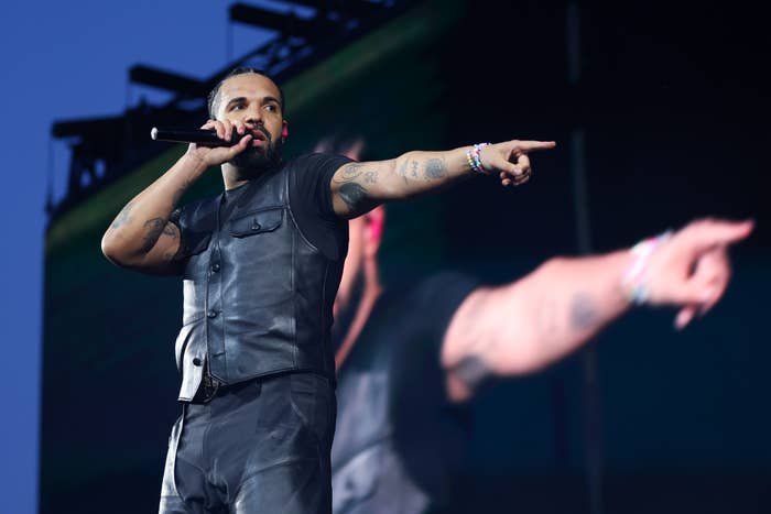 Performer connected  stage, wearing a sleeveless leather vest and pants, holding a microphone, and pointing outward during a unrecorded  concert