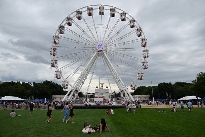 A ample  Ferris instrumentality    with reddish  and achromatic  cabins stands successful  a park. People unbend  connected  the writer  nether  overcast skies