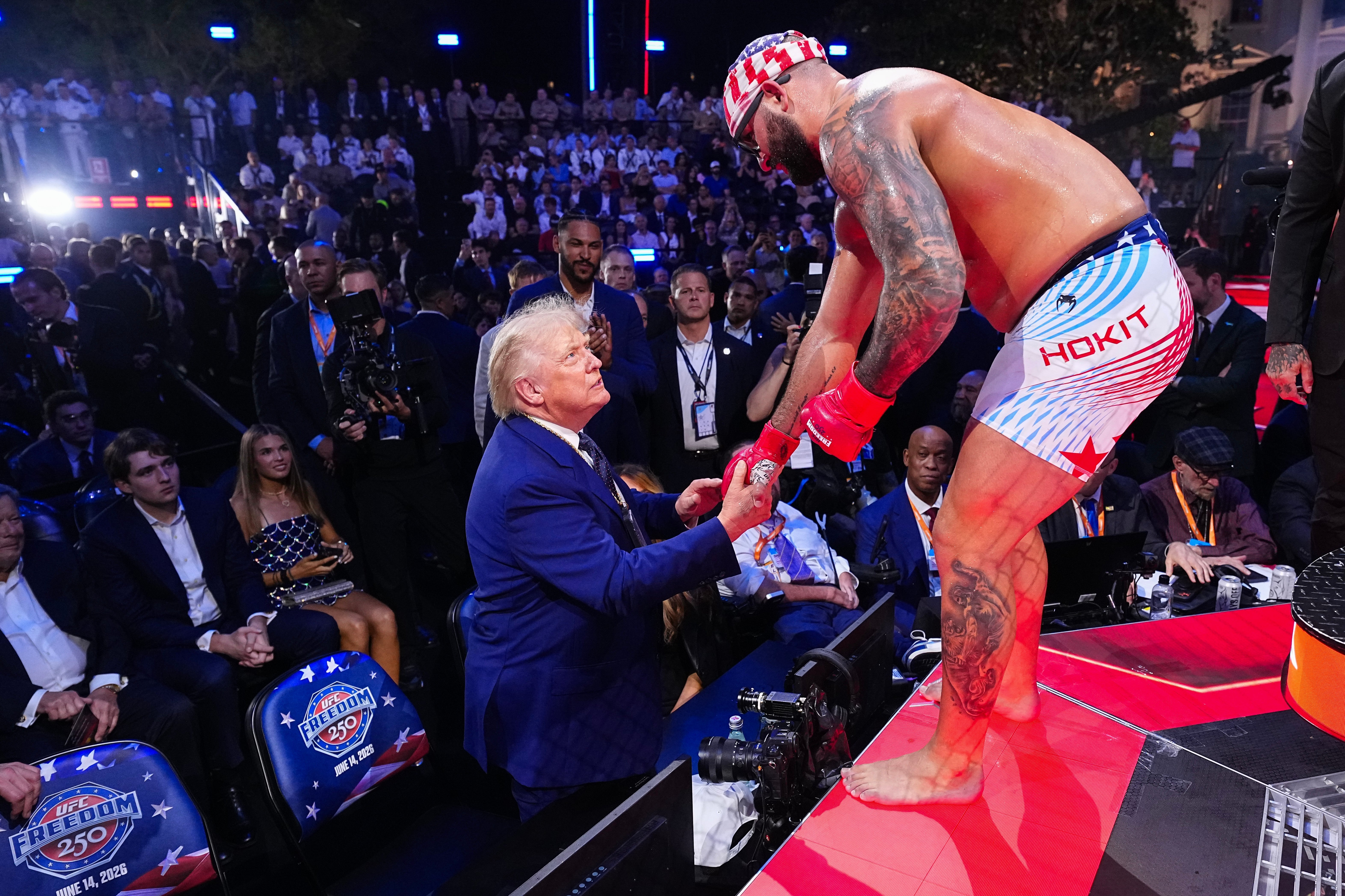 En mann i dress håndhilser på en MMA-fighter i shorts og caps på scenen blant en folkemengde på et arrangement