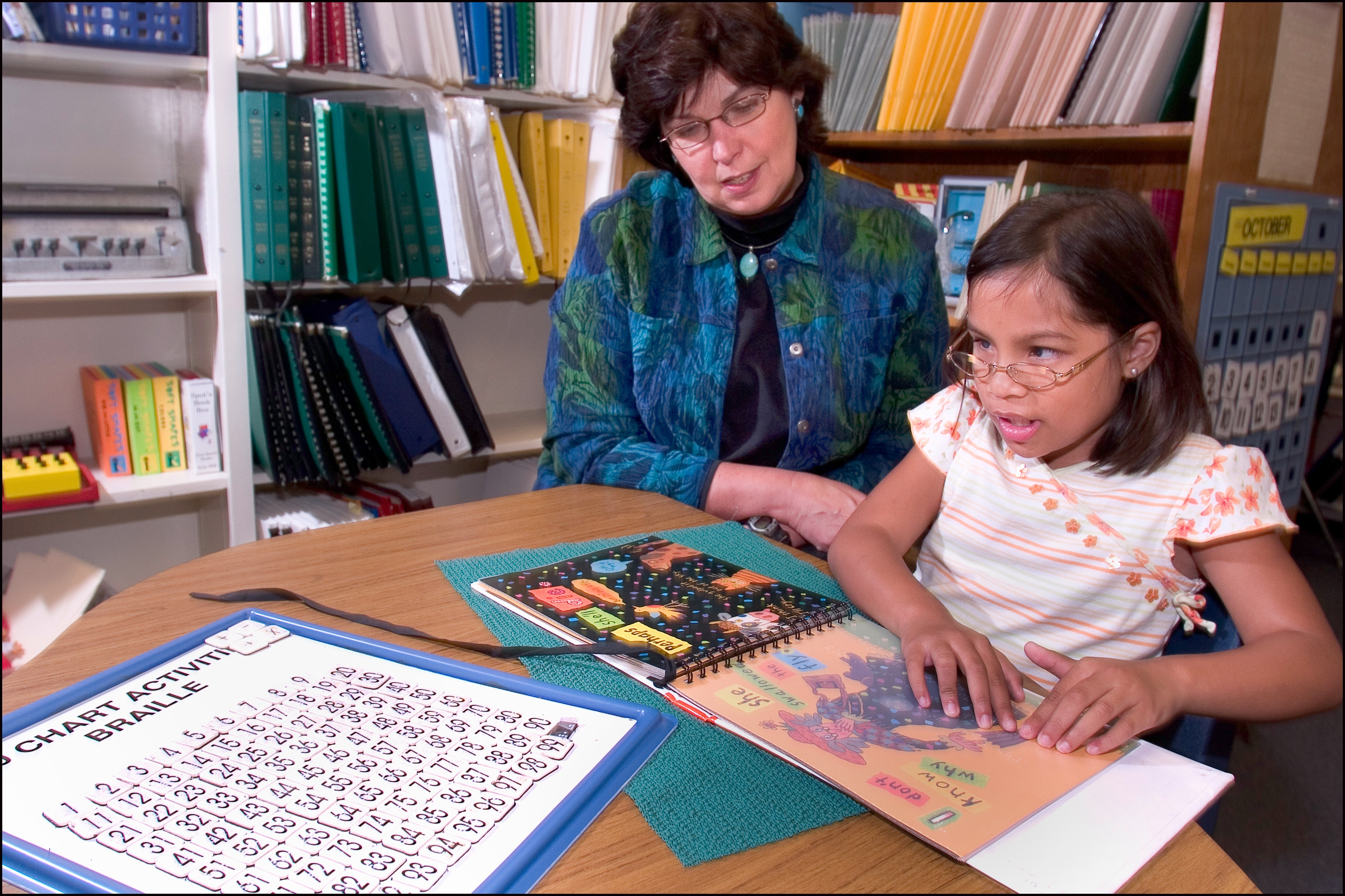 A young miss reads a Braille publication astatine a array with an pedagogue successful a room setting