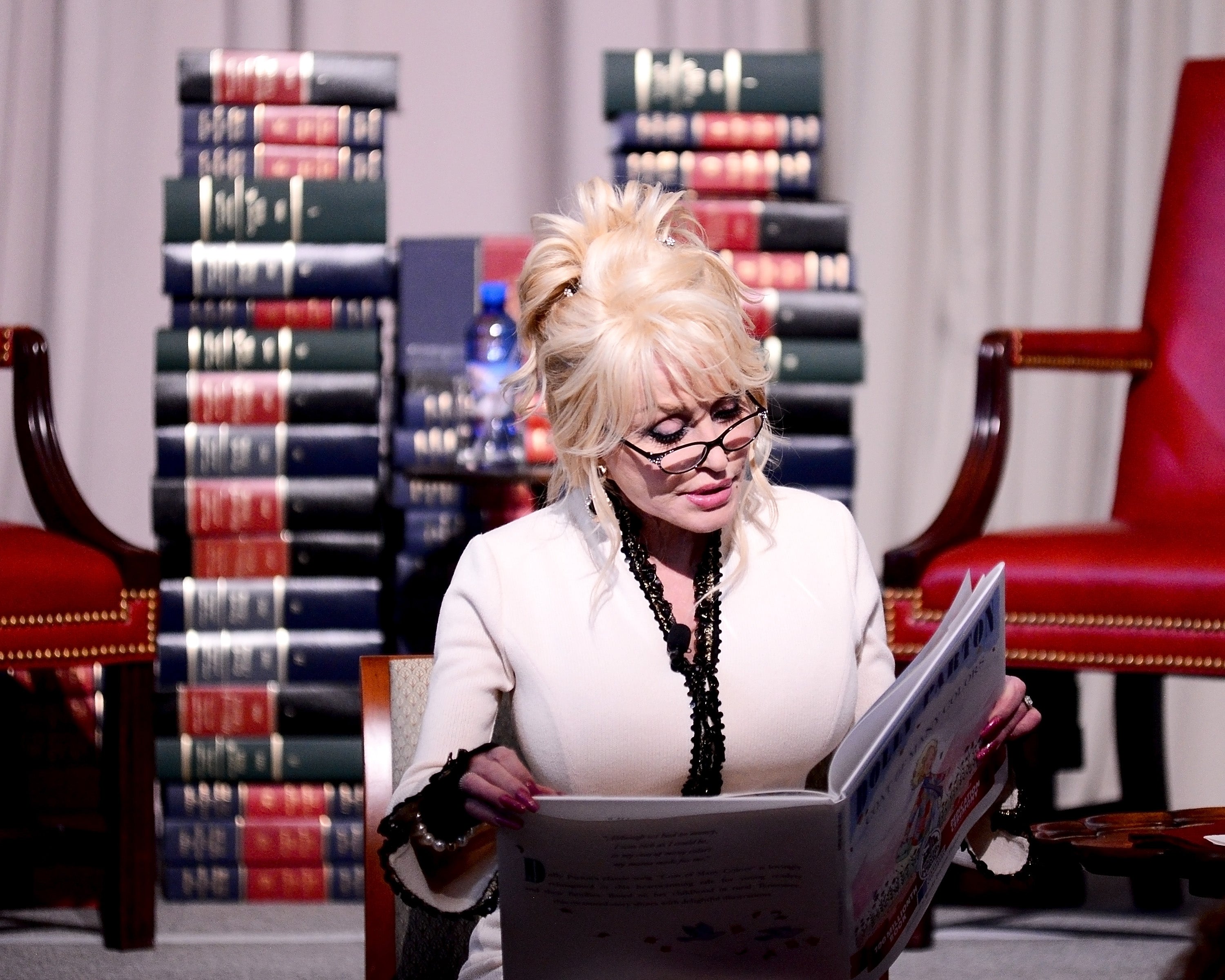 A idiosyncratic with glasses reads a book, sitting successful a chair. Stacks of books are successful the background