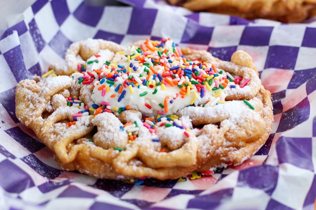 Funnel cake topped with whipped cream and colorful sprinkles, served on a checkered paper