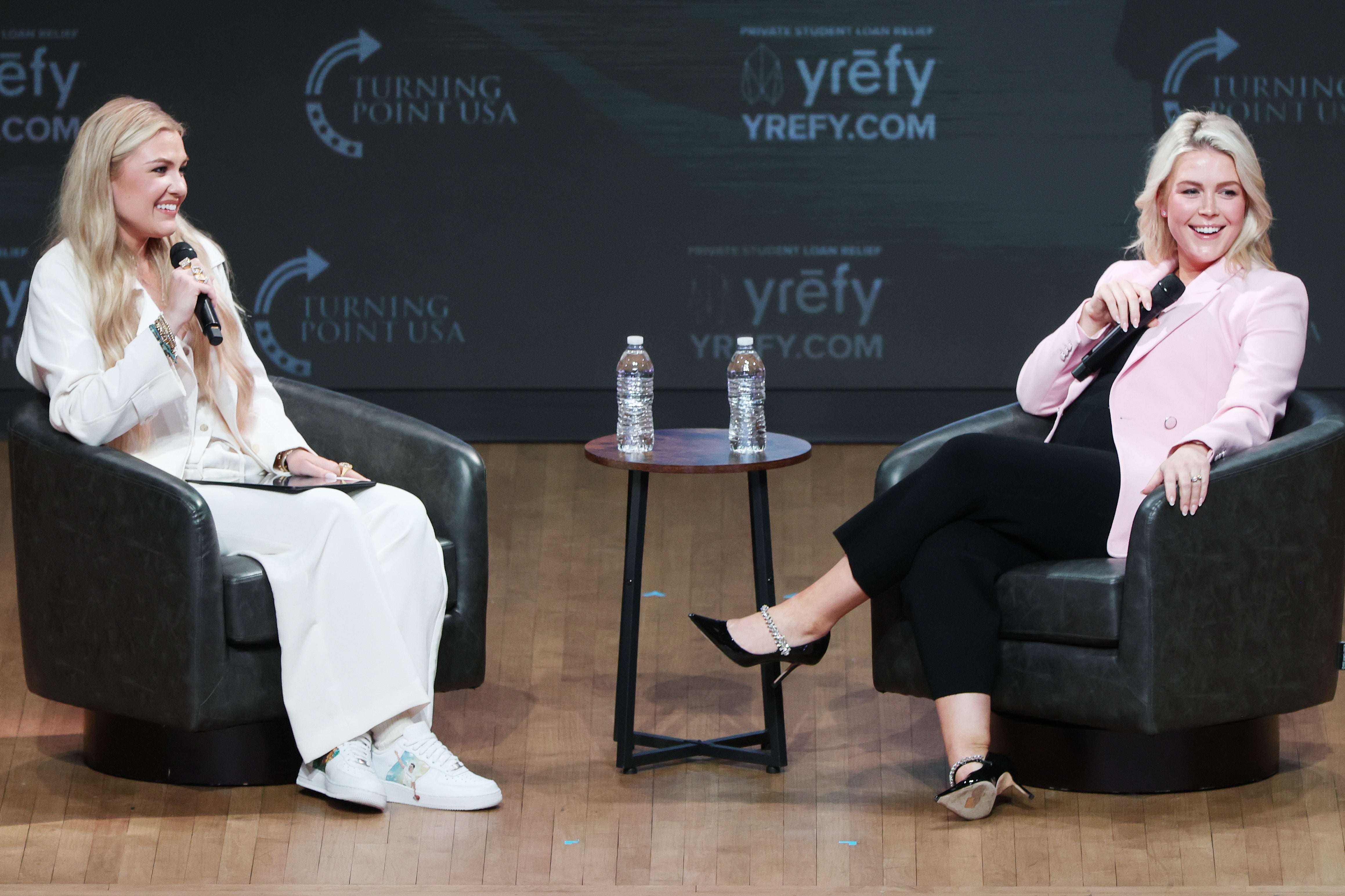 Two women beryllium   connected  signifier    successful  conversation. One wears a achromatic  outfit with sneakers, the different   a pinkish  blazer with achromatic  pants. Bottled h2o  connected  table