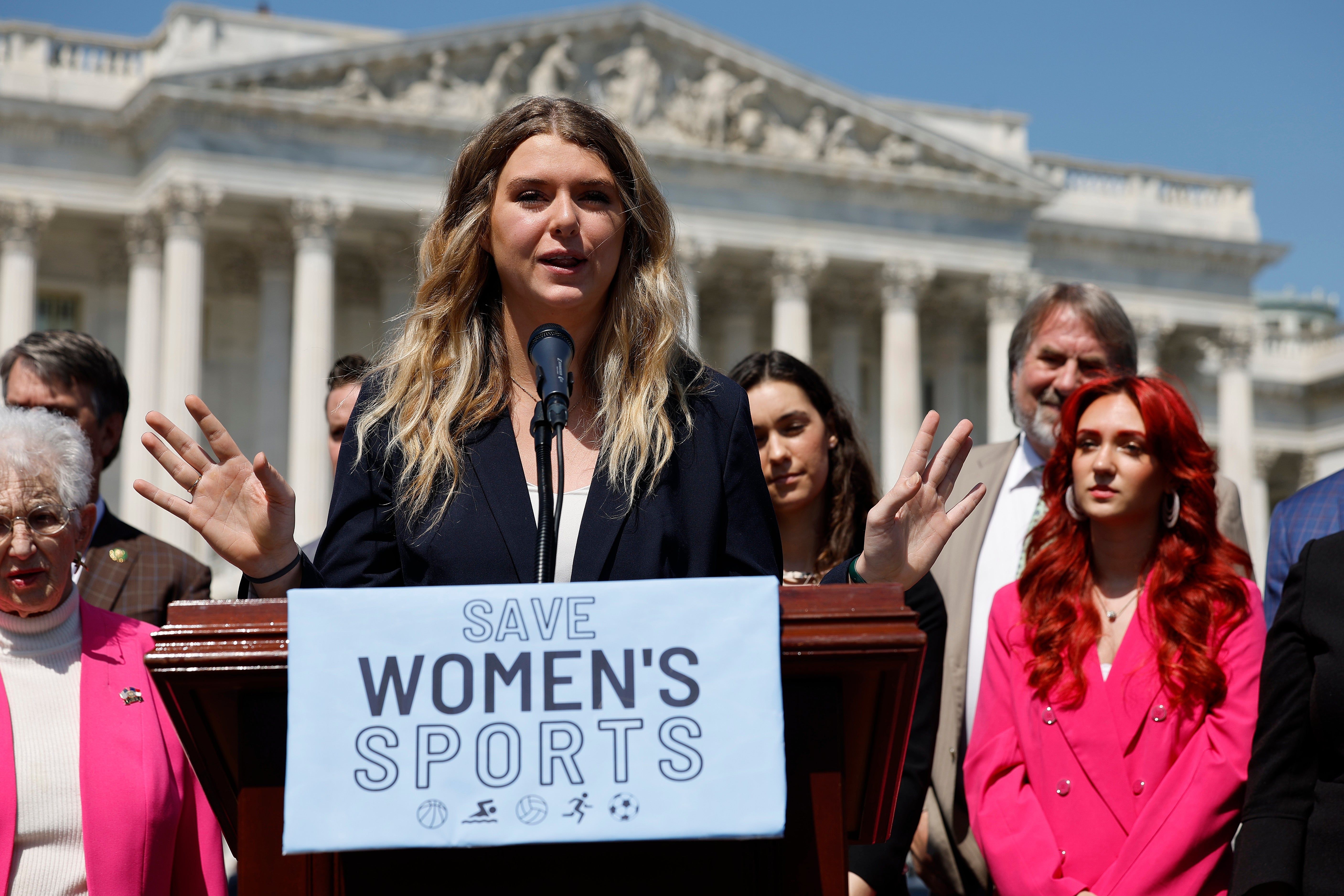 Person speaking astatine  a podium with "Save Women's Sports" sign, surrounded by a group. Building with columns successful  the background
