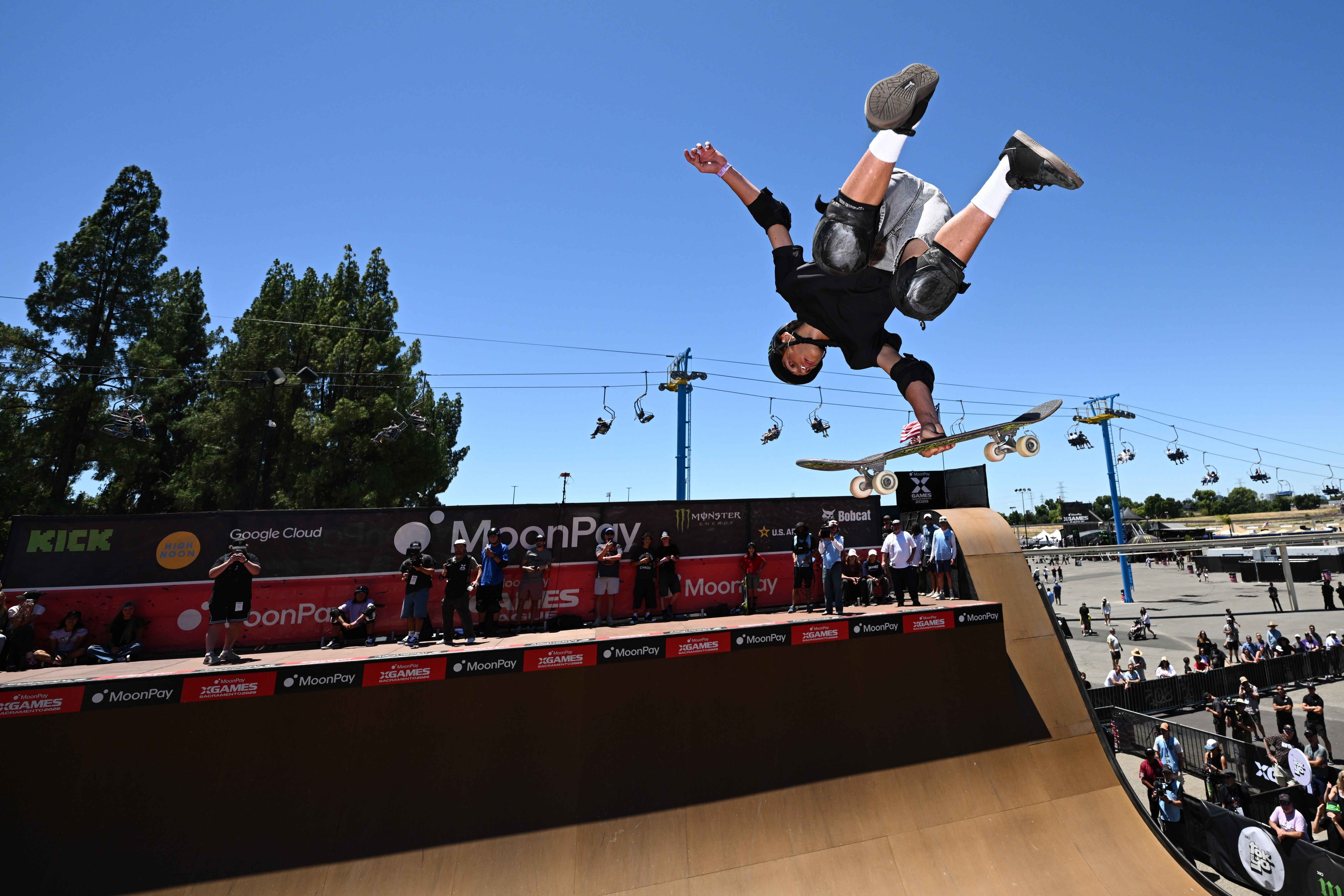 Skateboarder performs an aerial instrumentality   connected  a half-pipe astatine  an outdoor event, successful  mid-air with protective gear, portion    spectators watch