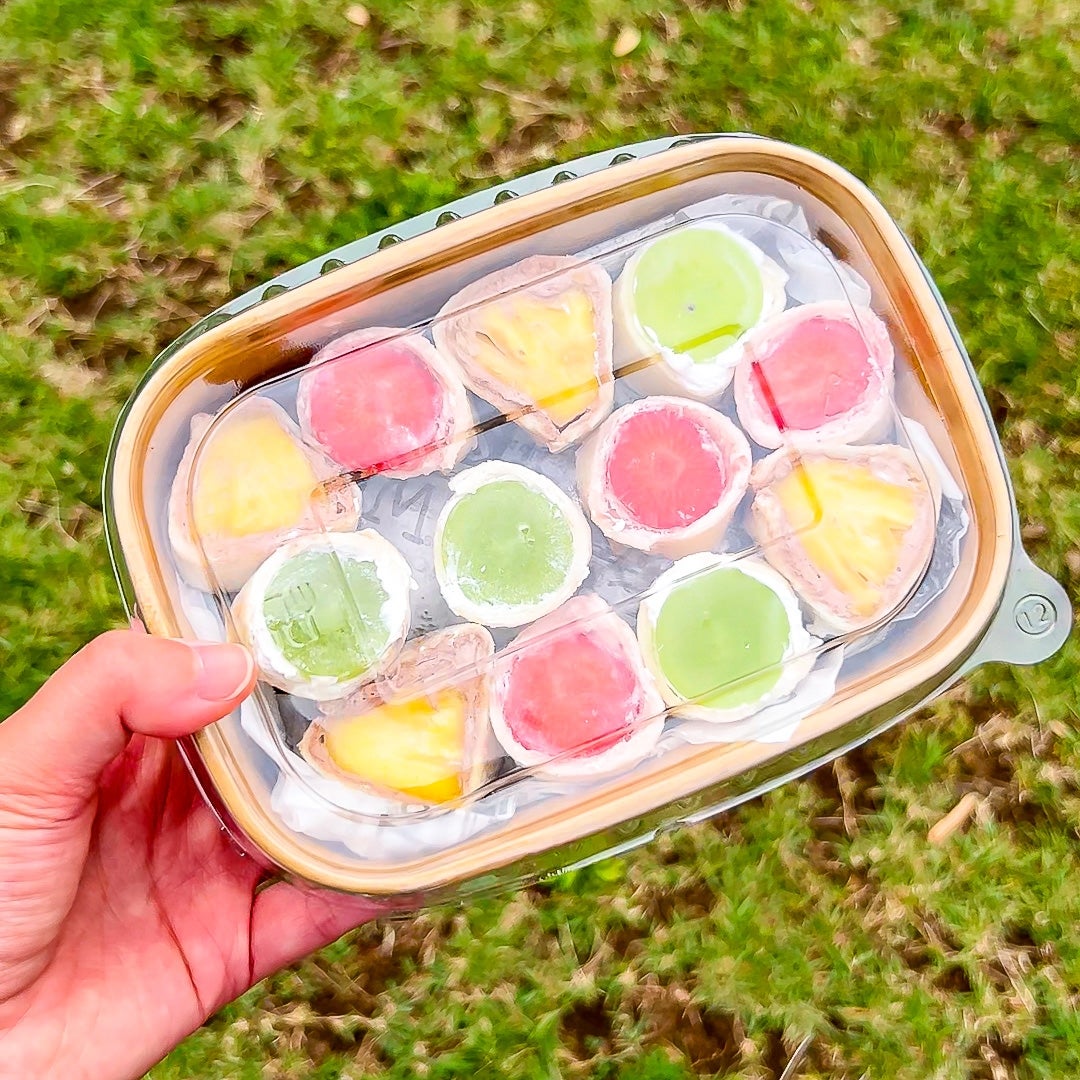 手に持たれた透明なプラスチック容器に入った12個の色とりどりの果物が巻かれたロールサンドイッチ。