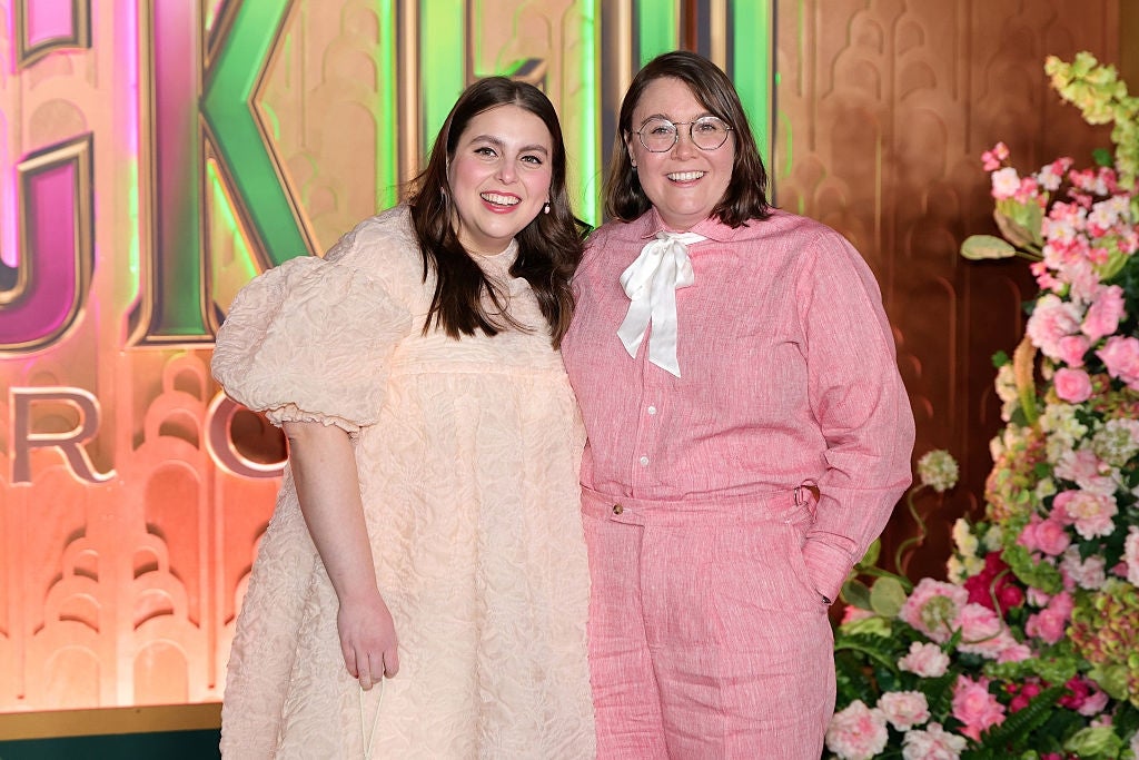 Two radical   smiling connected  a reddish  carpet; 1  wears a airy  dress, the different   a pinkish  suit   with a achromatic  bow. Background features flowers and decorative text