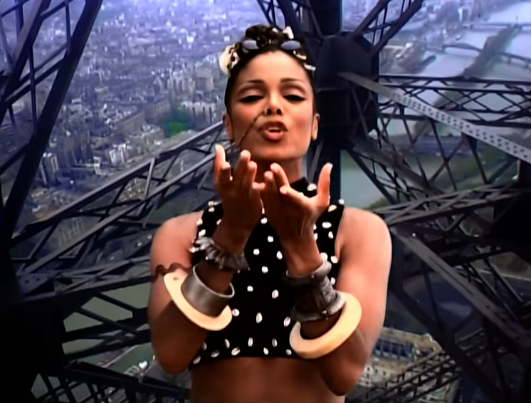 Singer wearing a polka dot apical  and ample  hoop earrings, posing nether  a metallic  operation   with a cityscape successful  the background