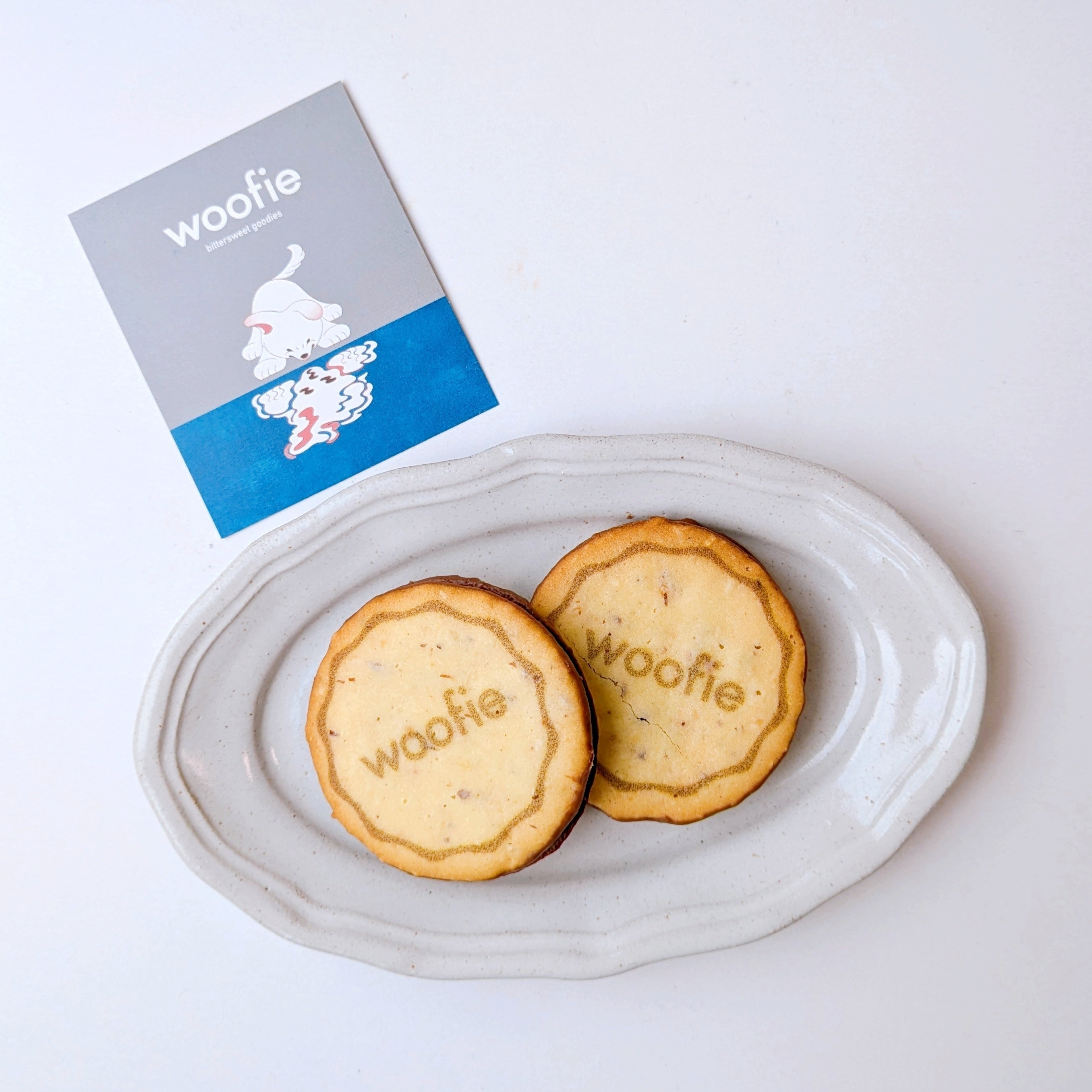 woofieの「サンドクッキー（キャラメル＆ヘーゼルナッツ）」が白いプレートに「woofie」と書かれたクッキー2枚が置かれ、横にロゴ入りのカードがあります。