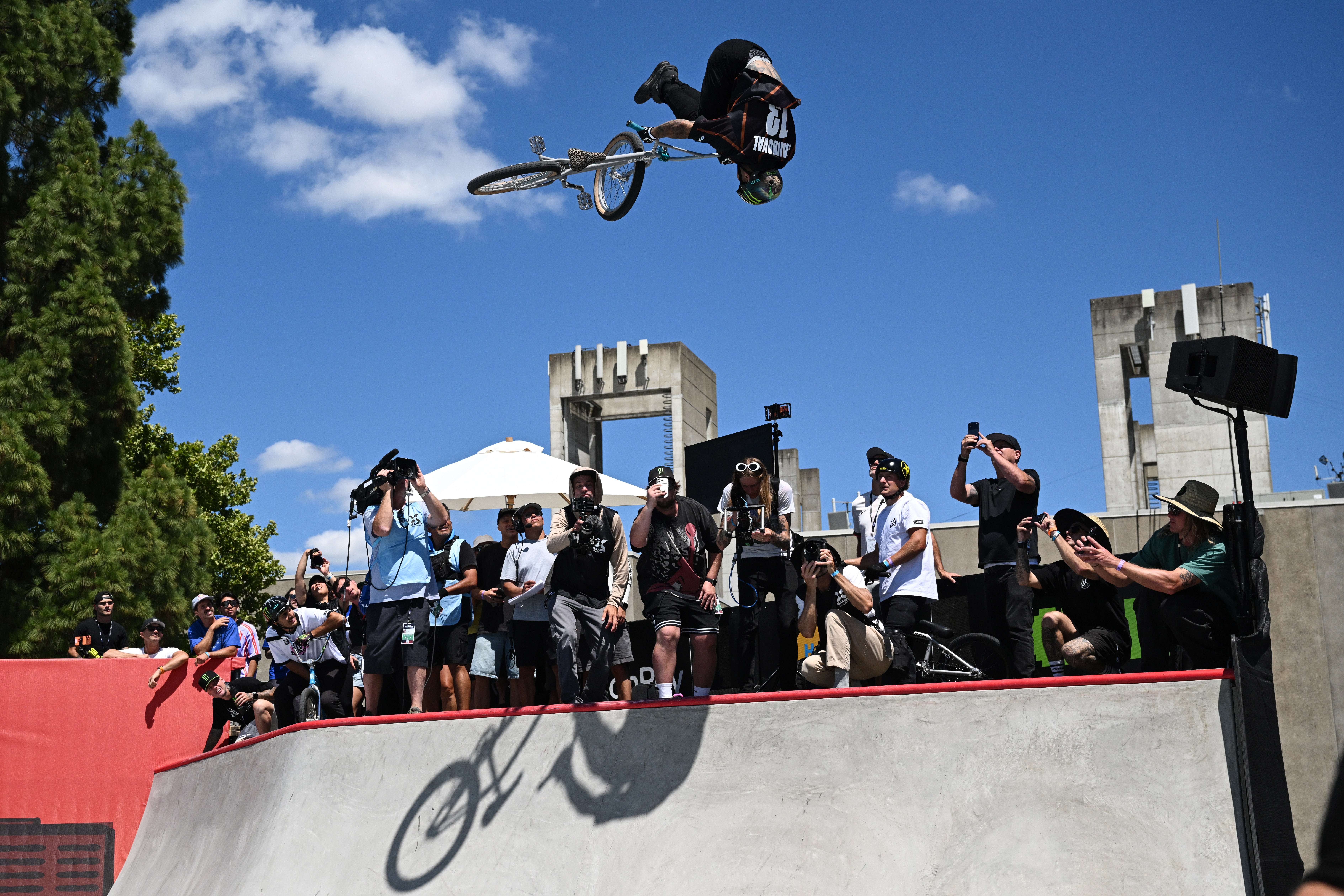 BMX rider performs an aerial instrumentality   supra  a skate parkland  ramp, watched by a assemblage  of spectators with cameras capturing the action