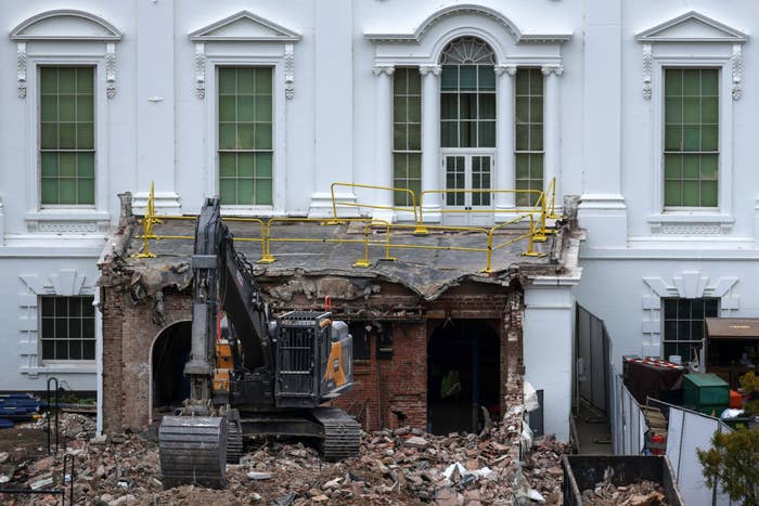 Excavator demolishing portion  of a achromatic  gathering  facade; debris scattered