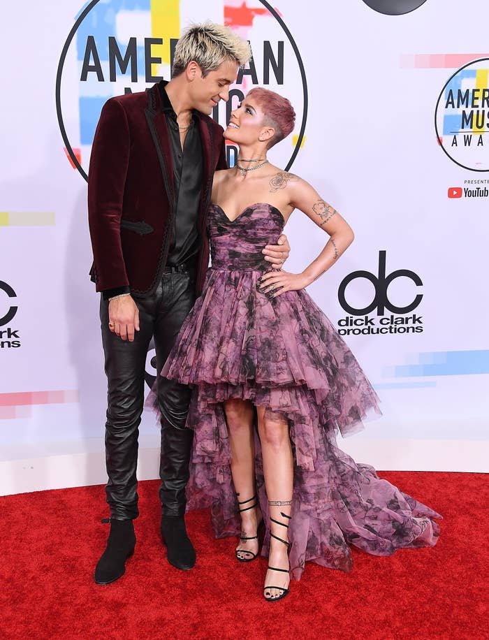 G-Eazy and Halsey on a red carpet: one in a velvet jacket and leather pants, the other in a strapless, layered gown and strappy heels, smiling at each other