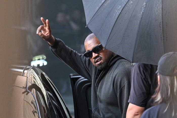 Kanye West in a hoodie and sunglasses makes a peace sign while getting out of a car under an umbrella