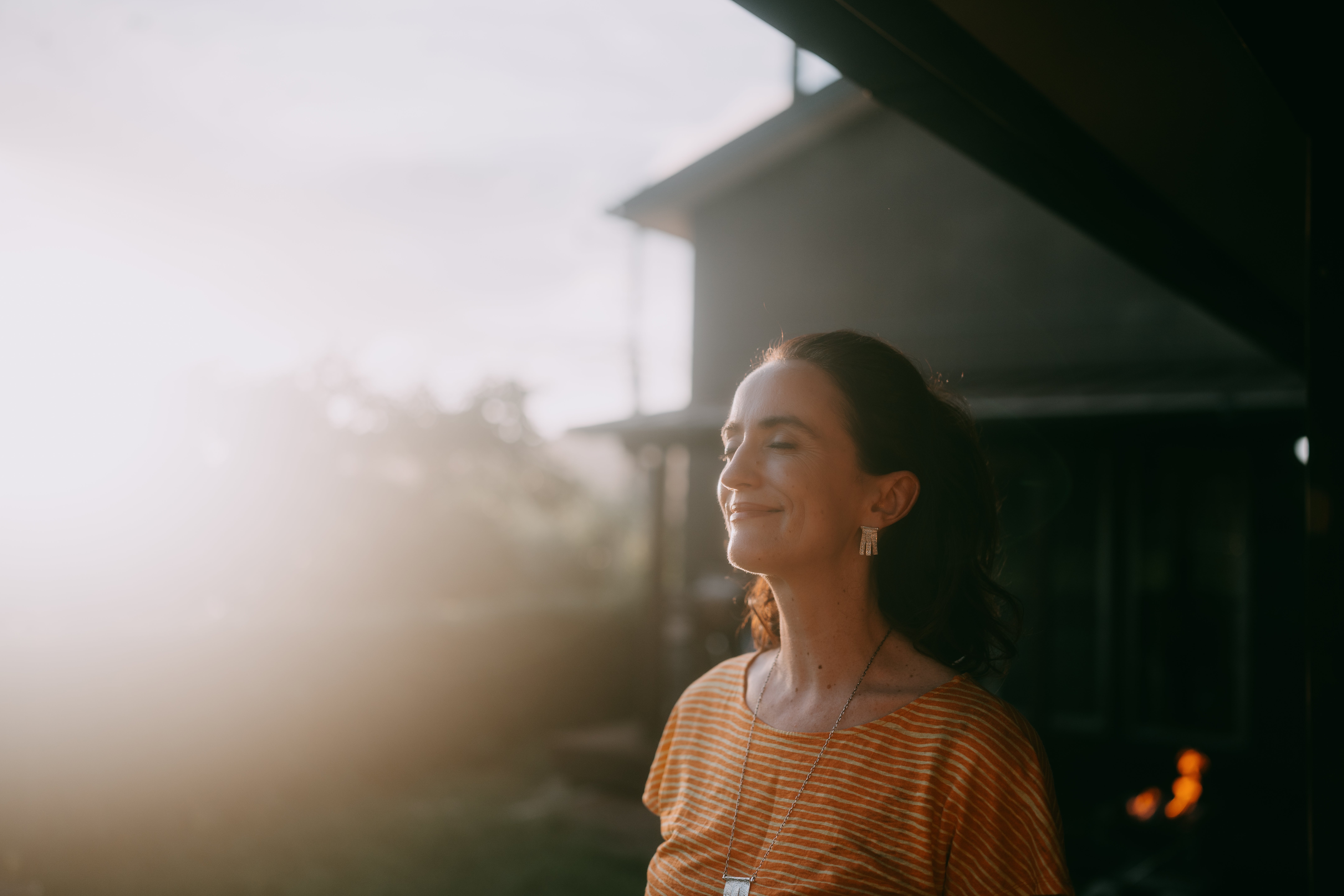 Person stands extracurricular  with eyes closed, smiling serenely successful  brushed  sunlight, wearing a casual apical  and jewelry, with a location   successful  the background