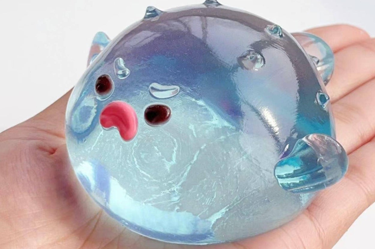 A hand holds a clear, round stress ball shaped like a pufferfish with an expressive face, ideal for fidgeting or relieving tension