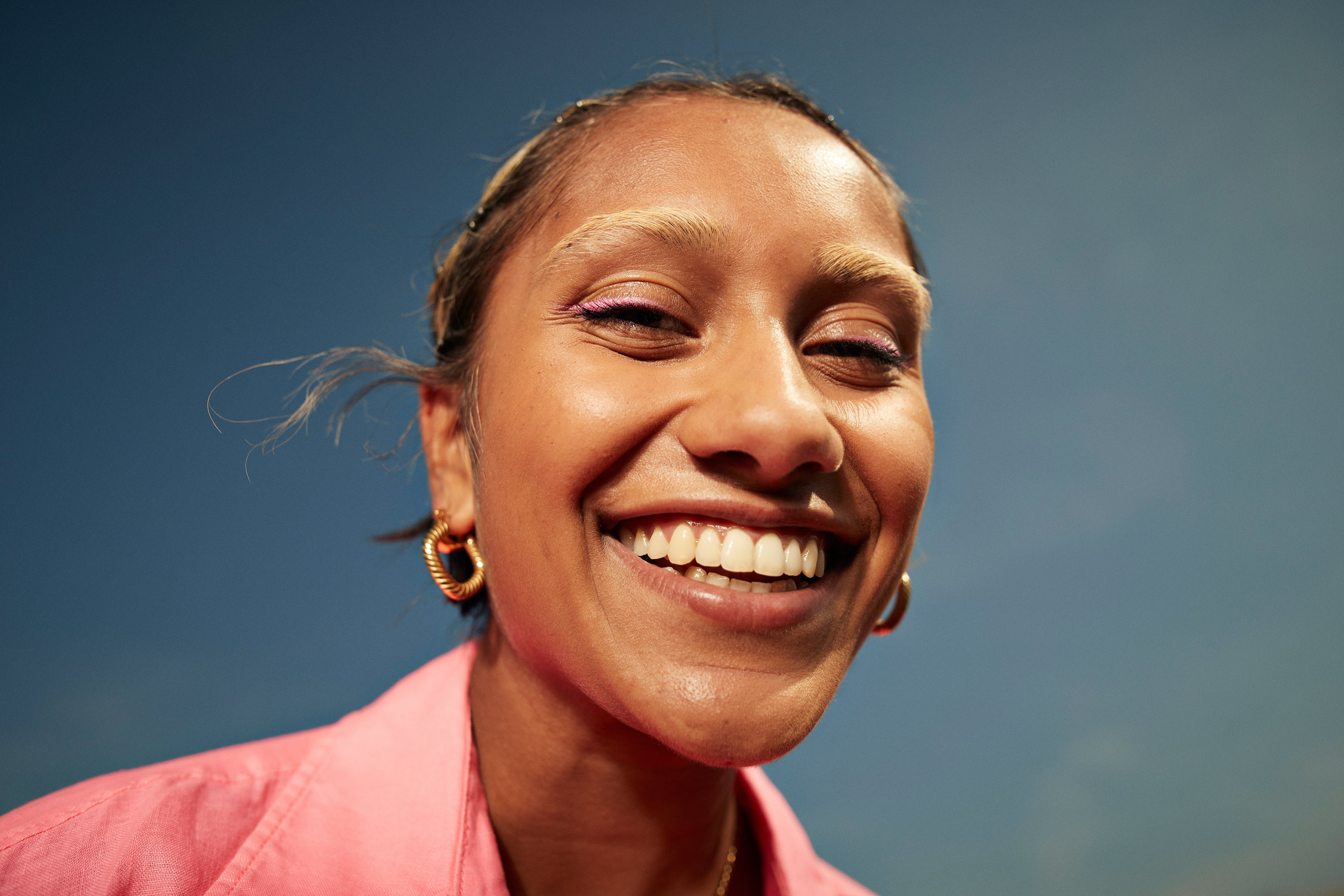 Person smiling wide against a wide sky, wearing hoop earrings and a pinkish shirt