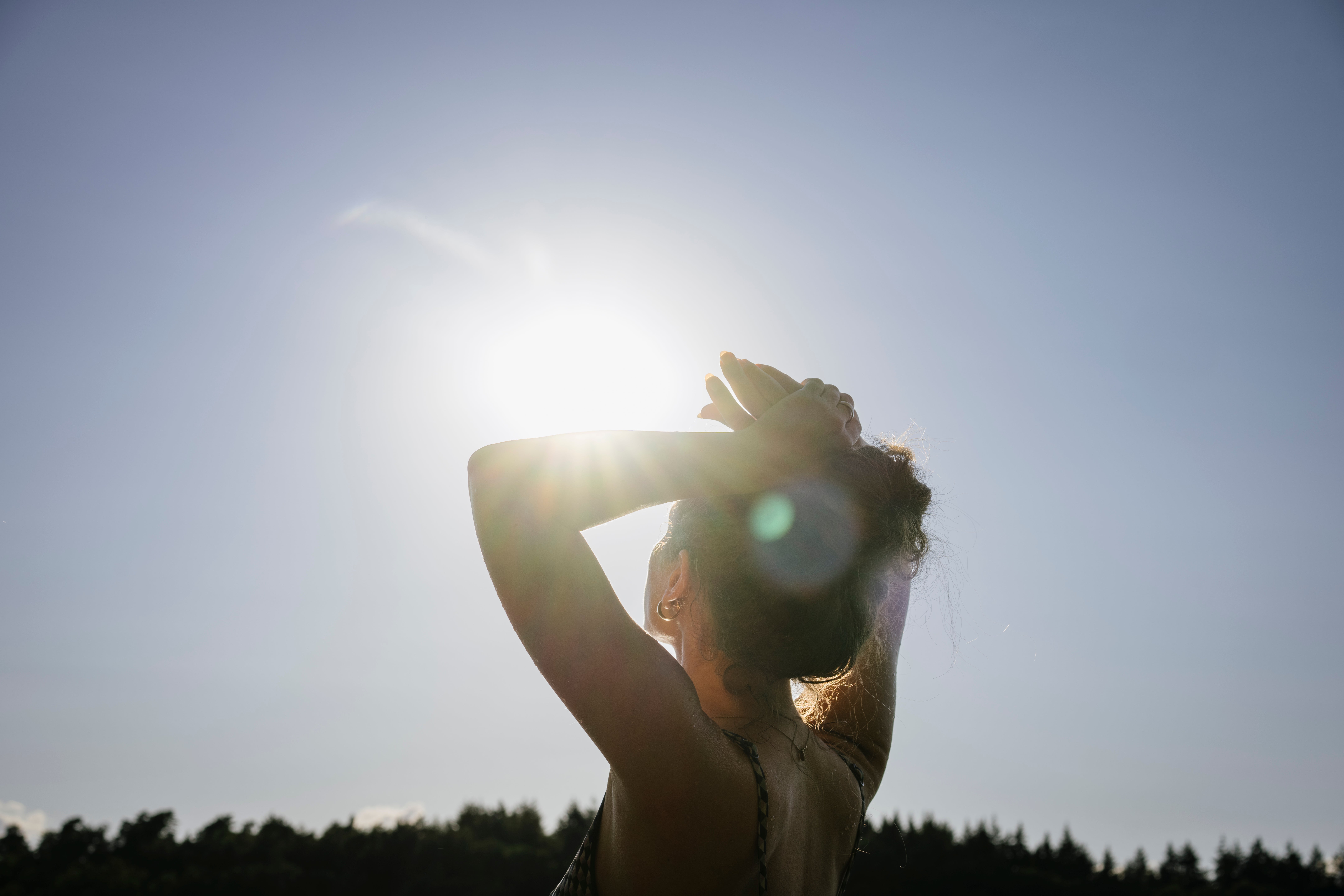 Person stands outdoors, silhouetted against the sky, hands lifted supra their head, with sunlight shining through