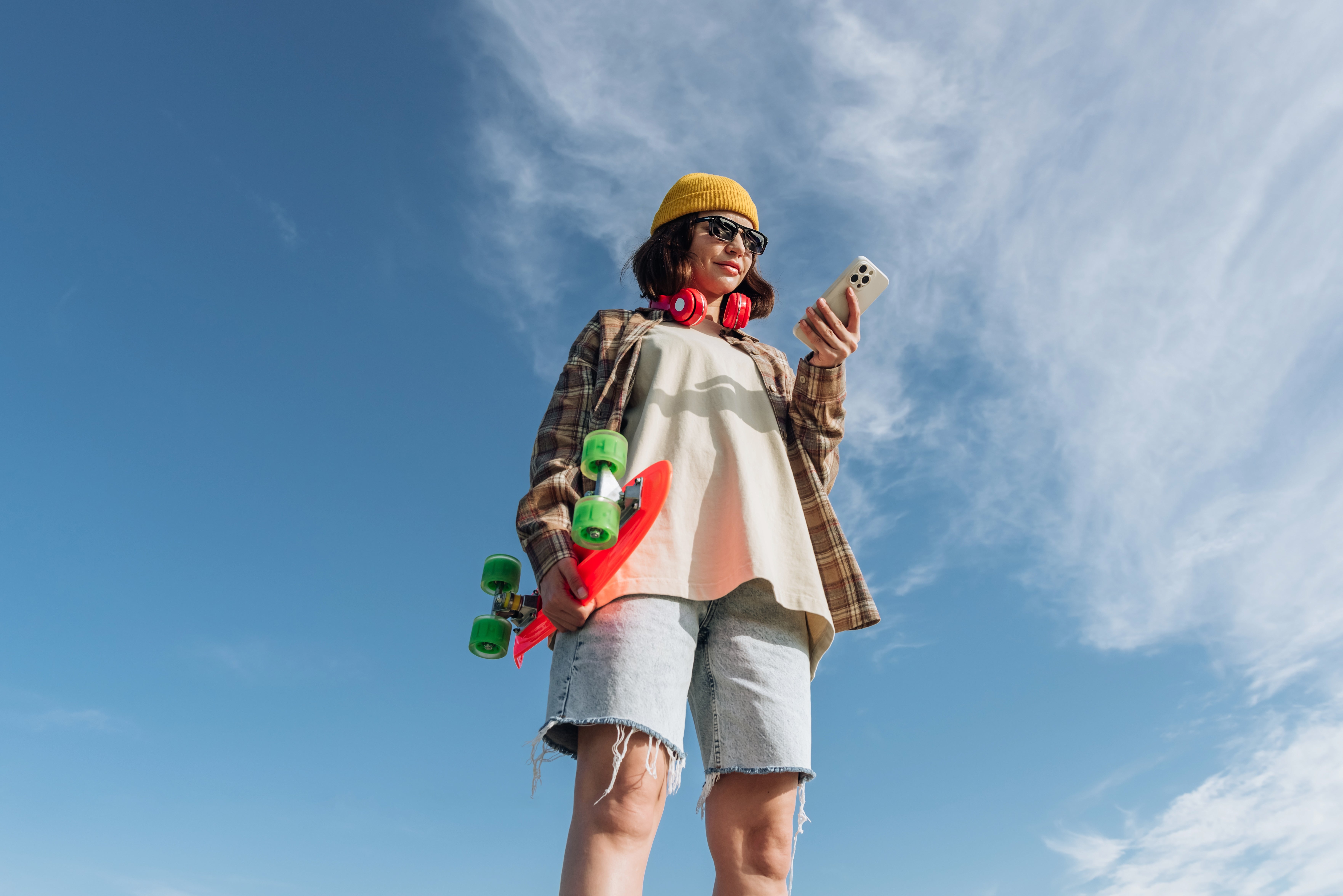 Person successful casual apparel with headphones, holding a skateboard and phone, stands against a bluish sky