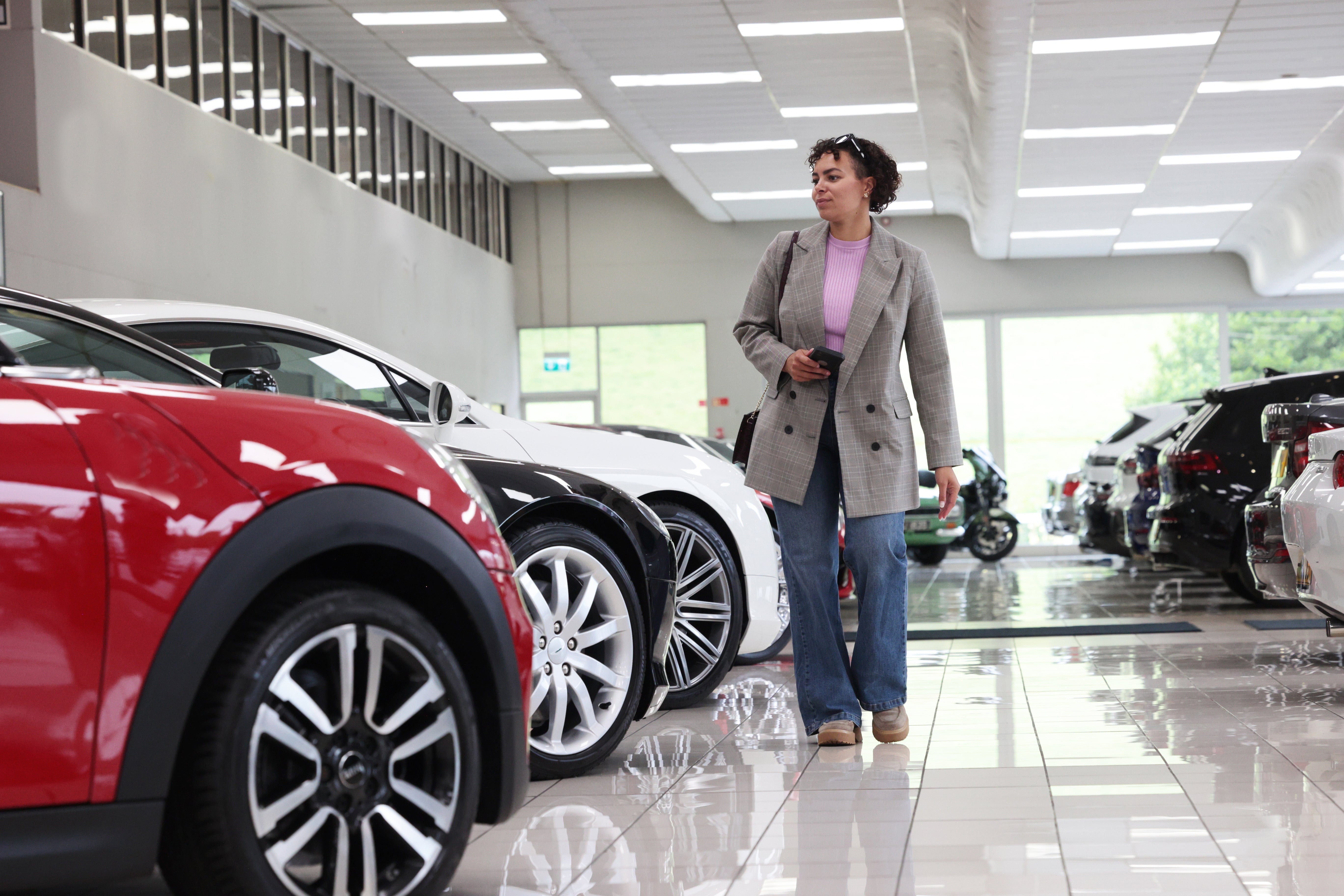 A idiosyncratic walks done a car showroom, looking astatine a enactment of parked vehicles. They deterioration a blazer, jeans, and clasp a smartphone