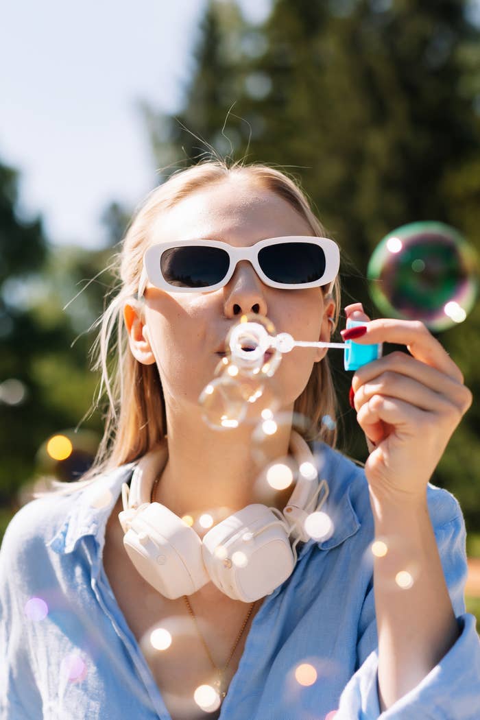 Person outdoors blowing bubbles, wearing sunglasses and headphones, successful  a casual shirt