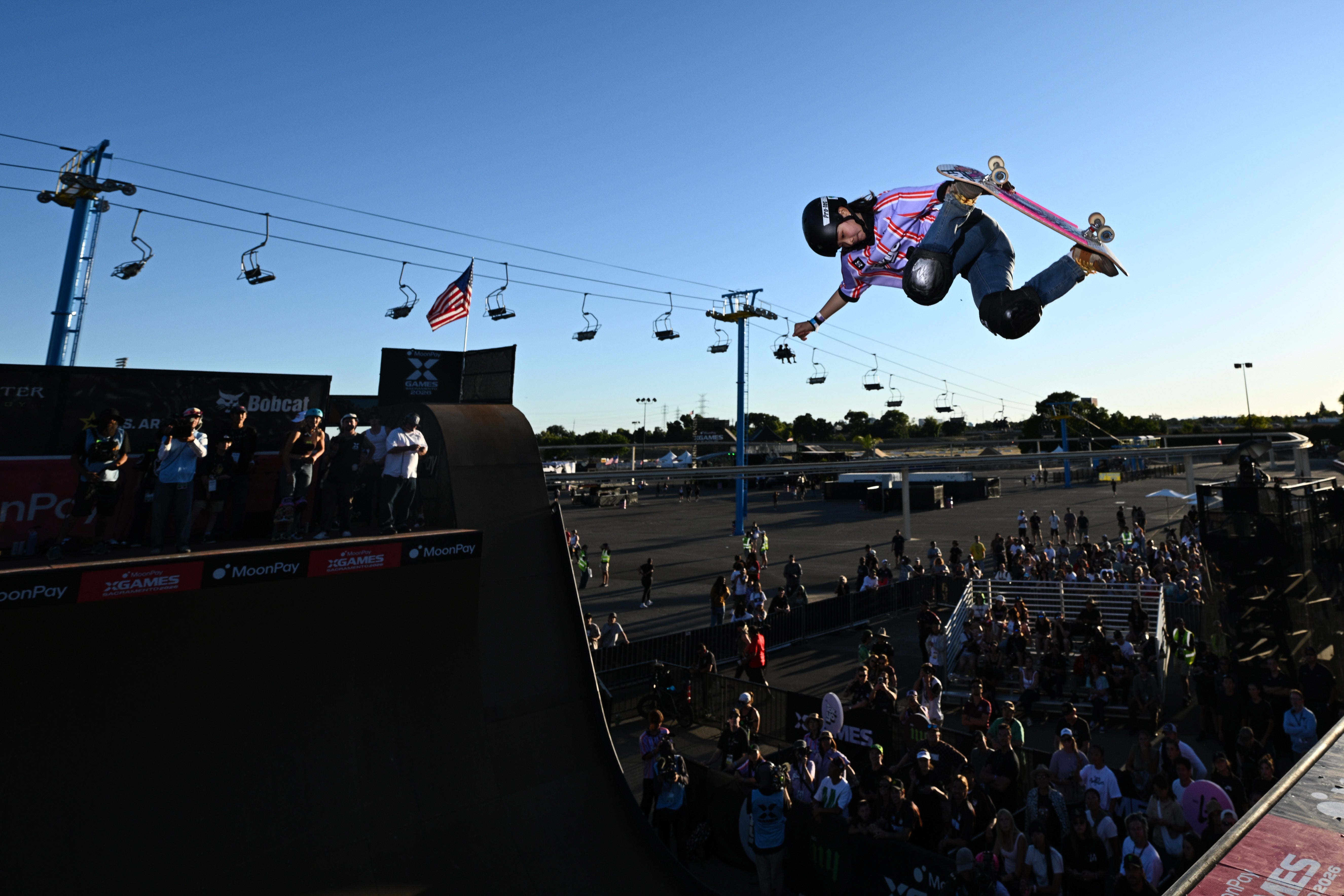 A skateboarder performs an aerial instrumentality   disconnected  a ramp with spectators watching, acceptable   against a entity  with skis  lifts successful  the background