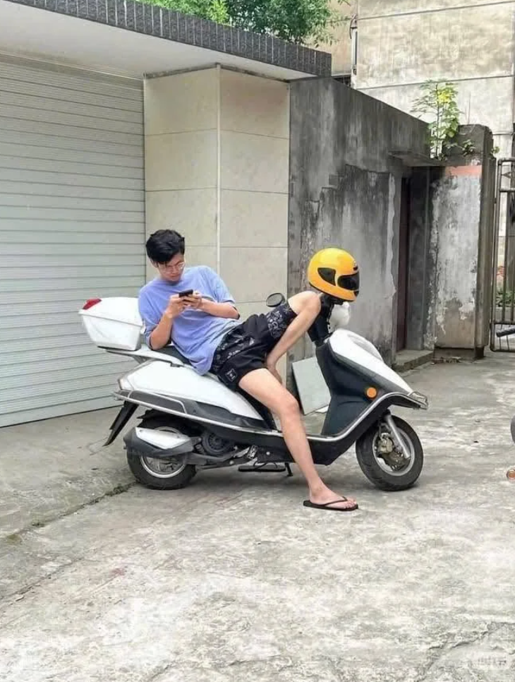 Person seated on a scooter, looking at a phone. The rider is dressed casually and another person is humorously sitting backwards on the scooter with a helmet on