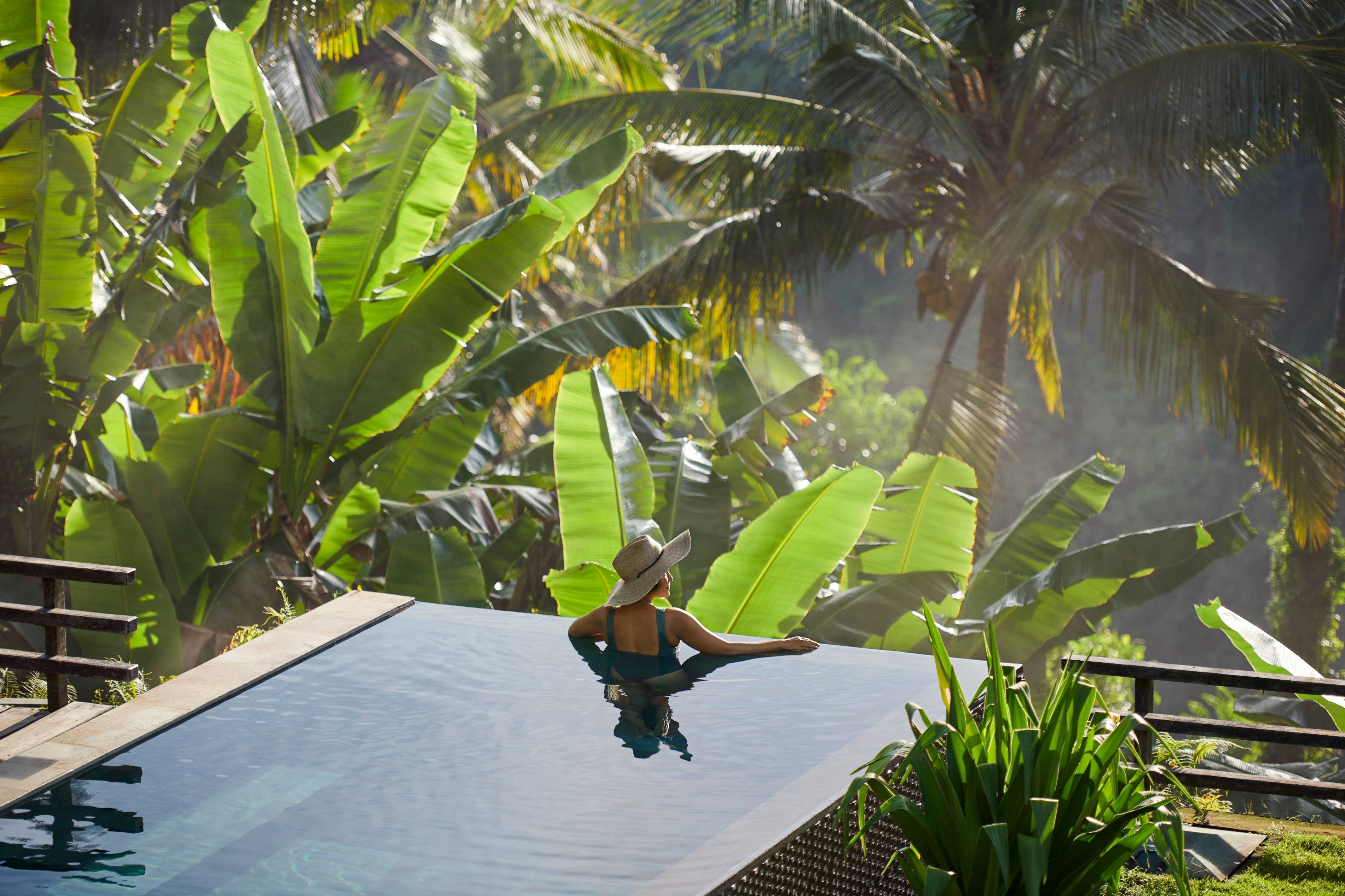 Person in a hat relaxing at an infinity pool, surrounded by lush tropical plants