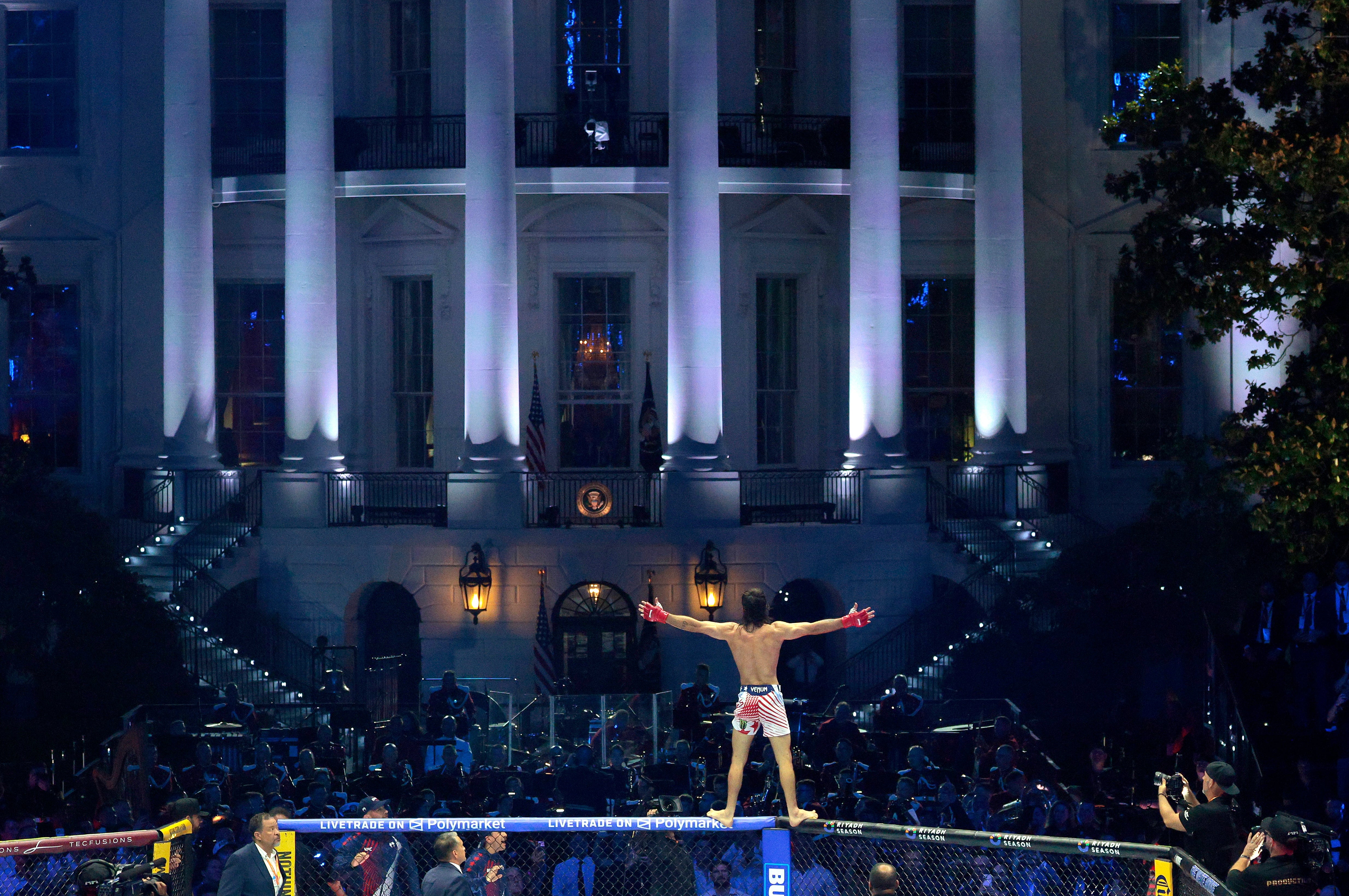 A idiosyncratic stands connected an MMA cage with arms outstretched successful beforehand of a large, illuminated gathering astatine night. Spectators and media situation the scene