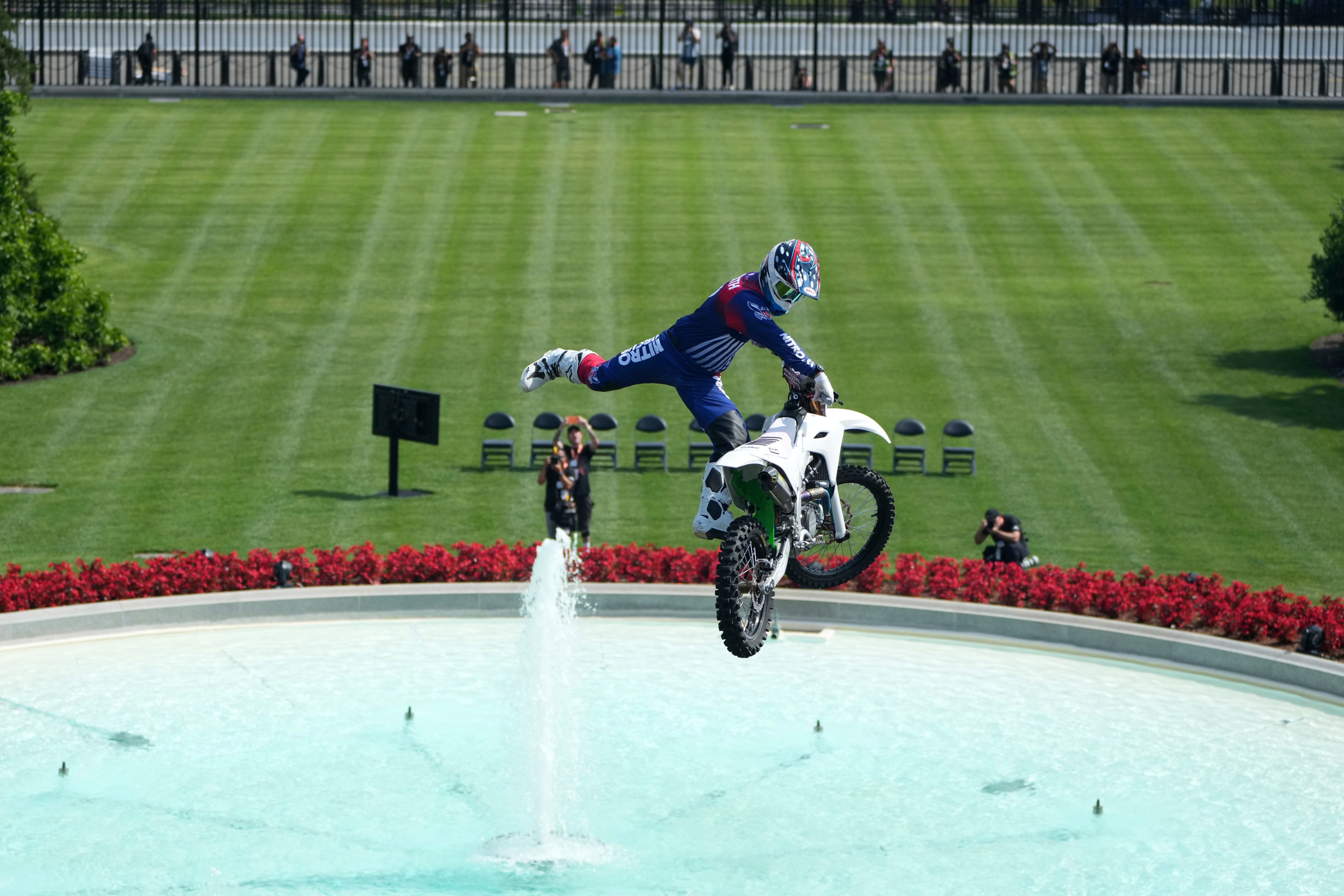 Motorcyclist performs a mid-air stunt supra a fountain with spectators and photographers successful the background