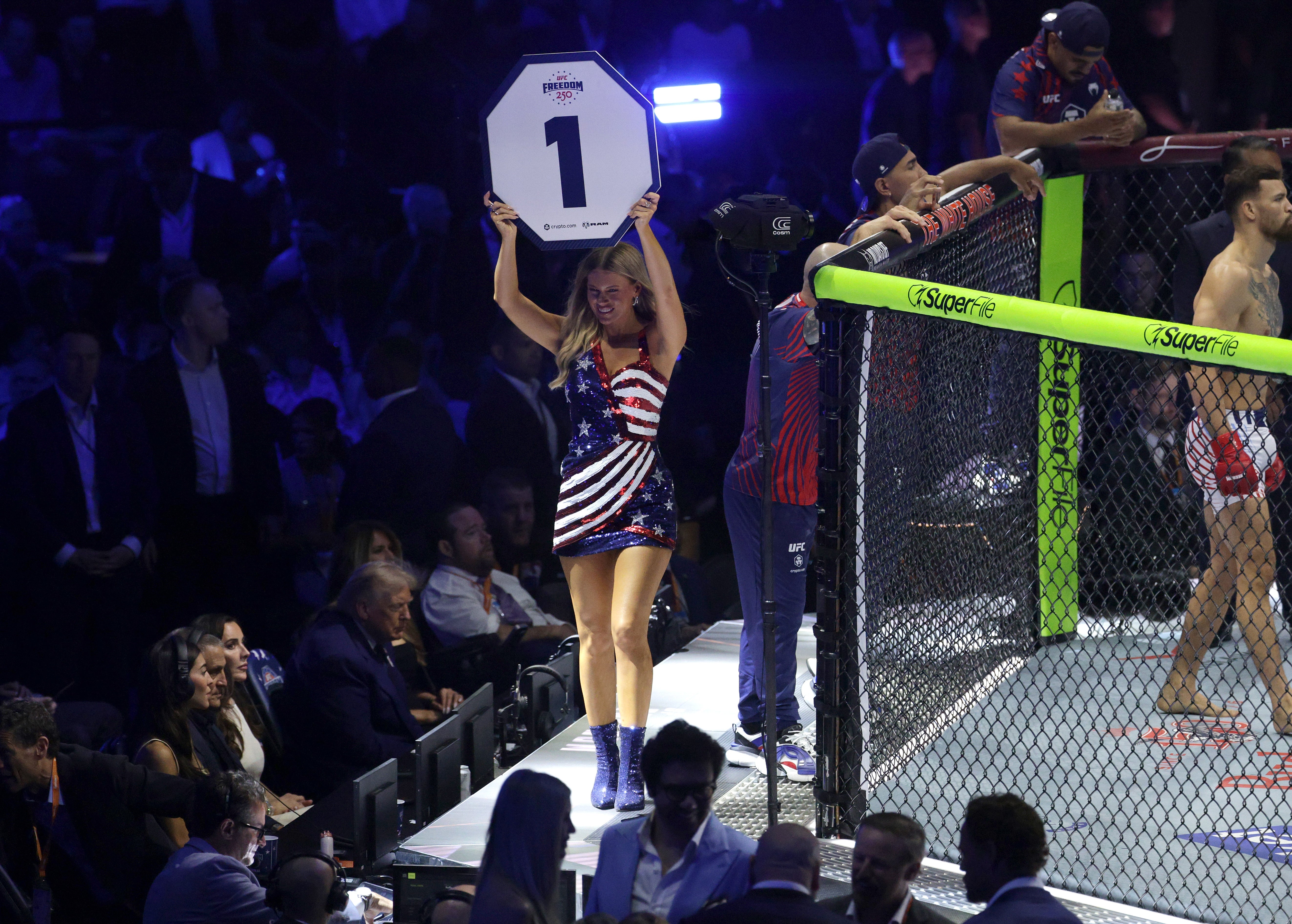 MMA lawsuit octagon, ringing miss holds "Round 1" sign; combatant squad stands by the cage. Patriotic-themed formal and boots. Spectators visible