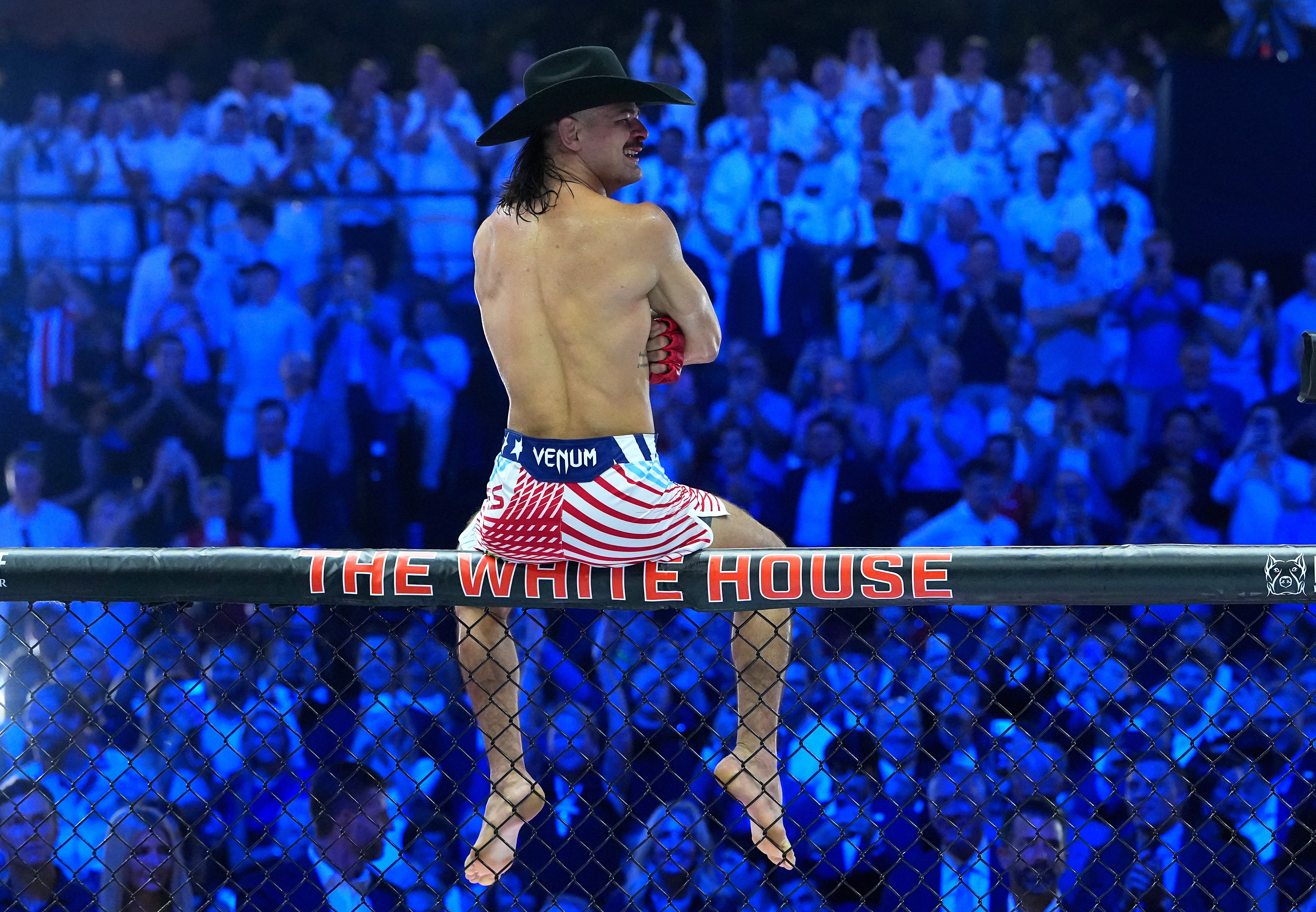 Person successful a cowboy chapeau and USA-themed diversion attire sitting connected an MMA cage with "The White House" text, surrounded by a cheering crowd