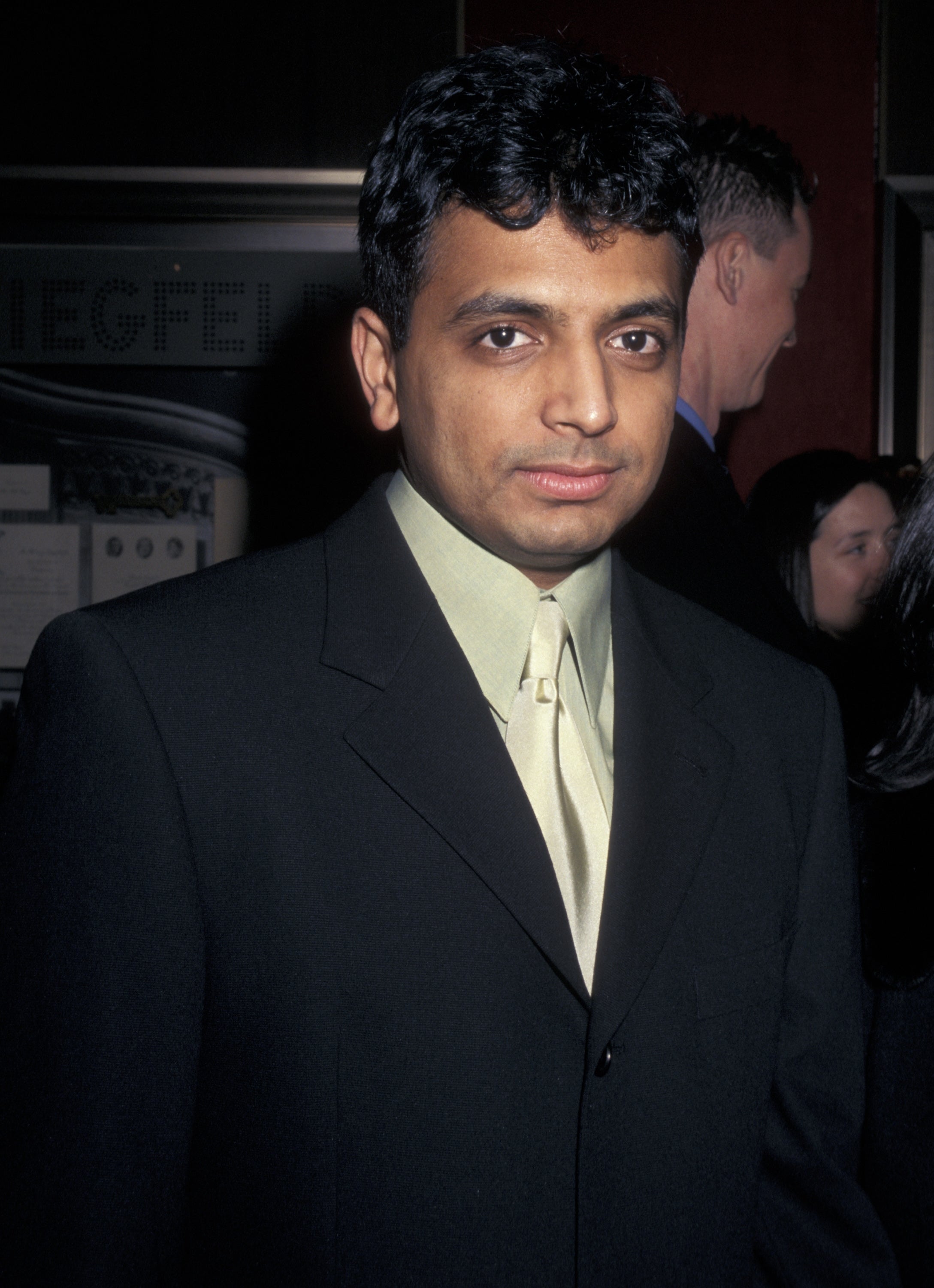 M. Night Shyamalan wearing a ceremonial  suit   stands astatine  an event, appearing poised and attentive