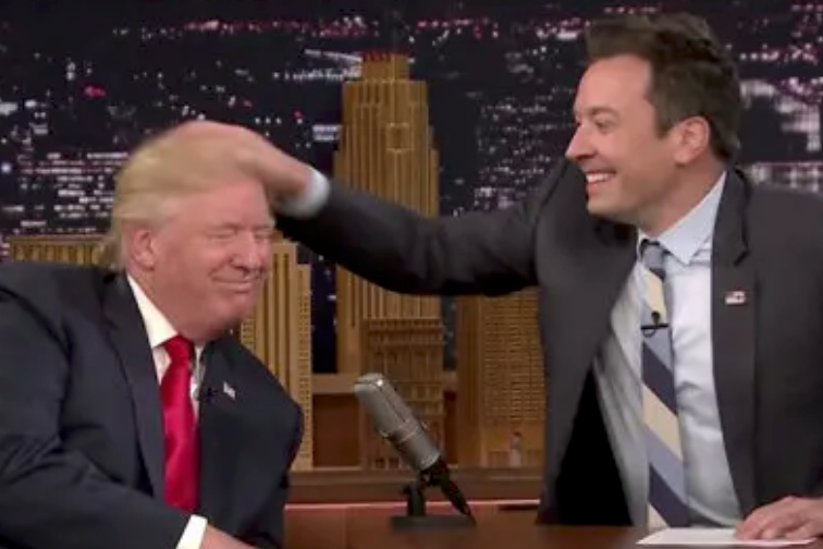 A talk show host playfully ruffles a guest's hair during an interview. Both are seated with a cityscape backdrop