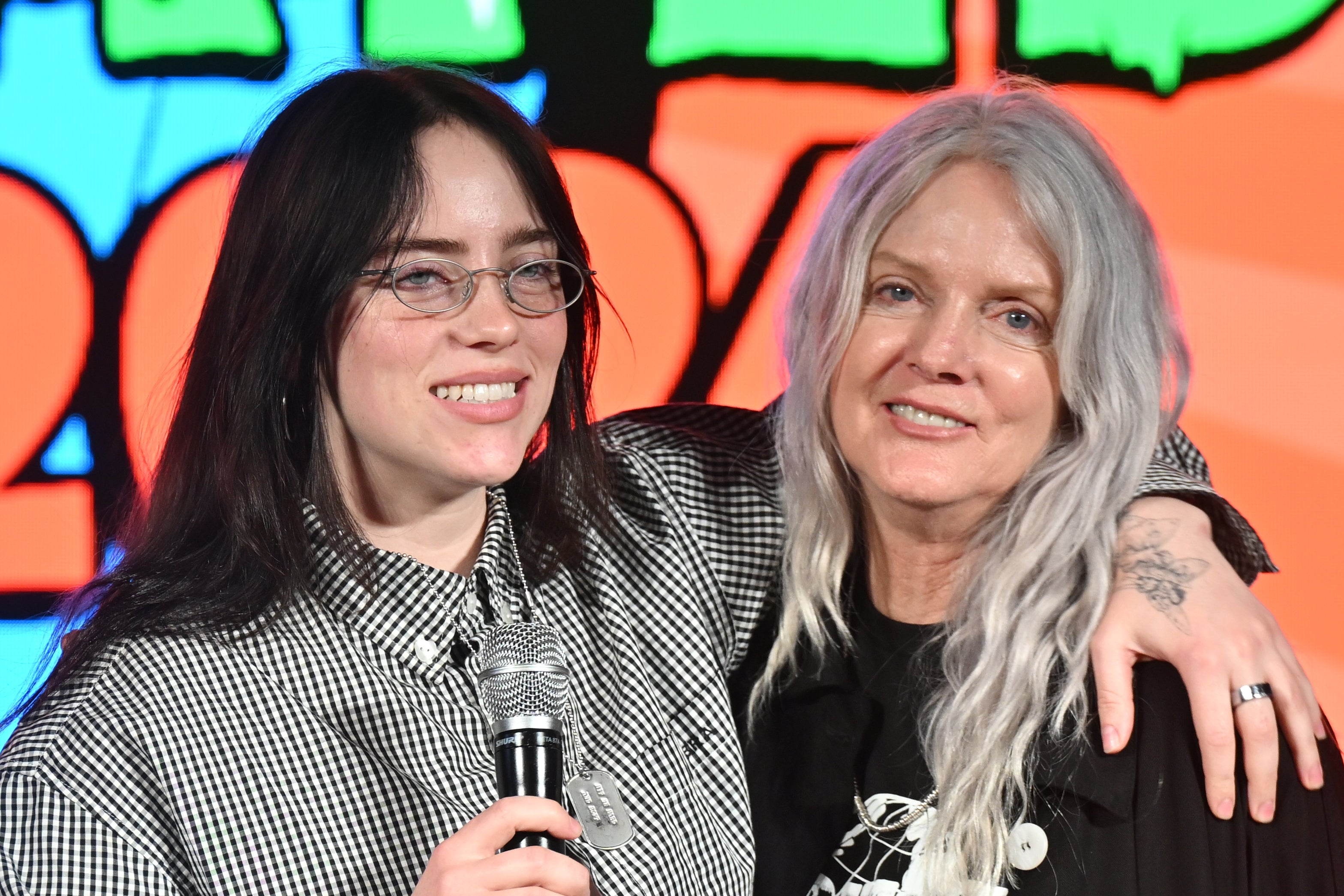Two people smiling at an event. One holds a microphone and wears plaid; the other wears a graphic tee and long coat. They have their arms around each other