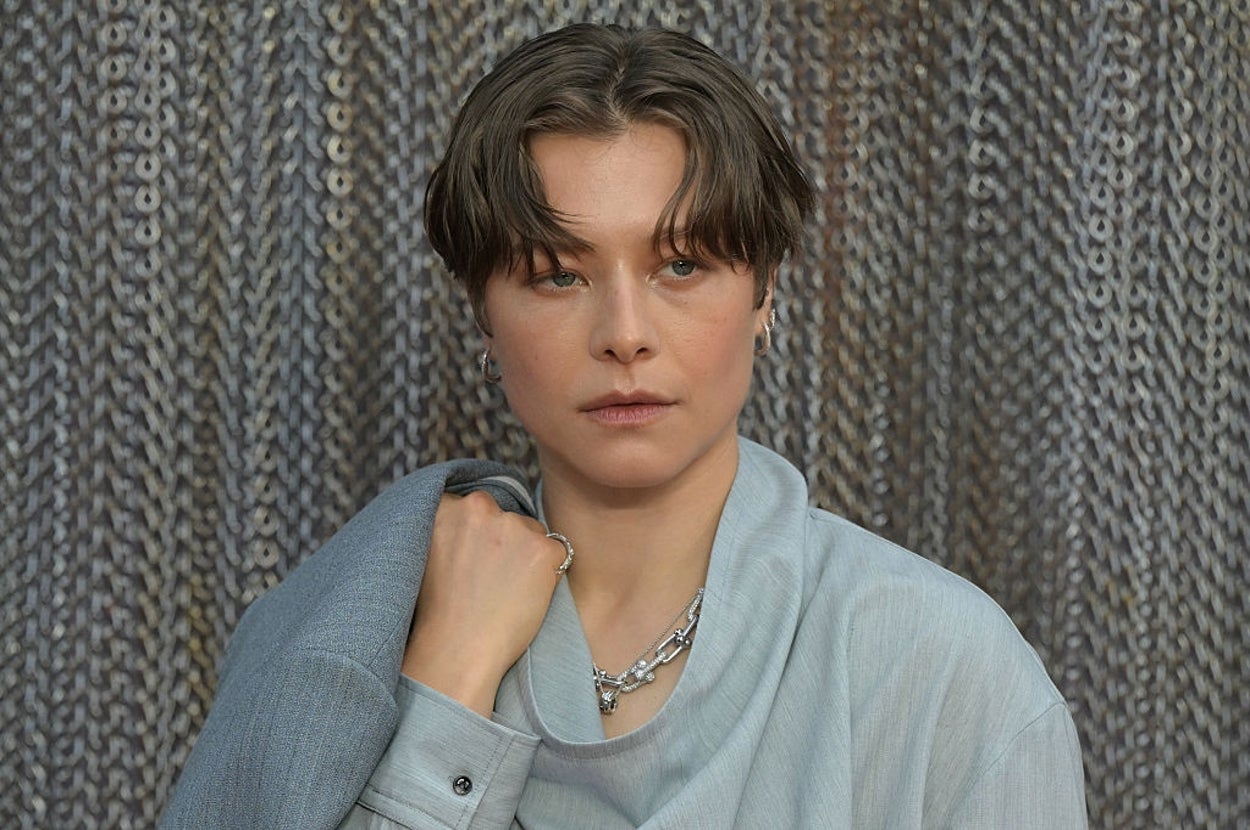 Person in draped suit with short hair poses in front of a textured backdrop, looking thoughtfully to the side