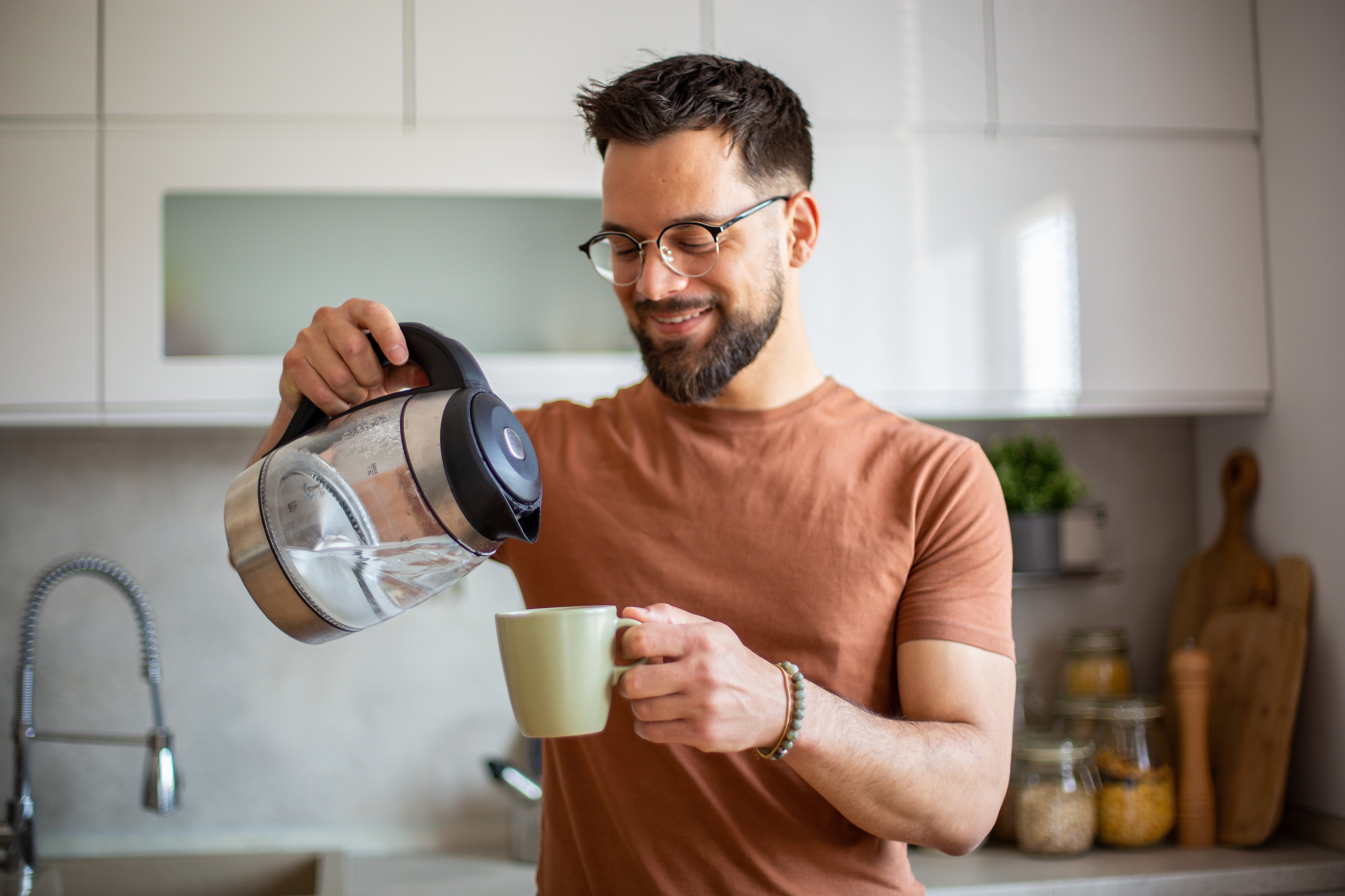 Man with glasses smiling while pouring coffee into a mug in a kitchen setting