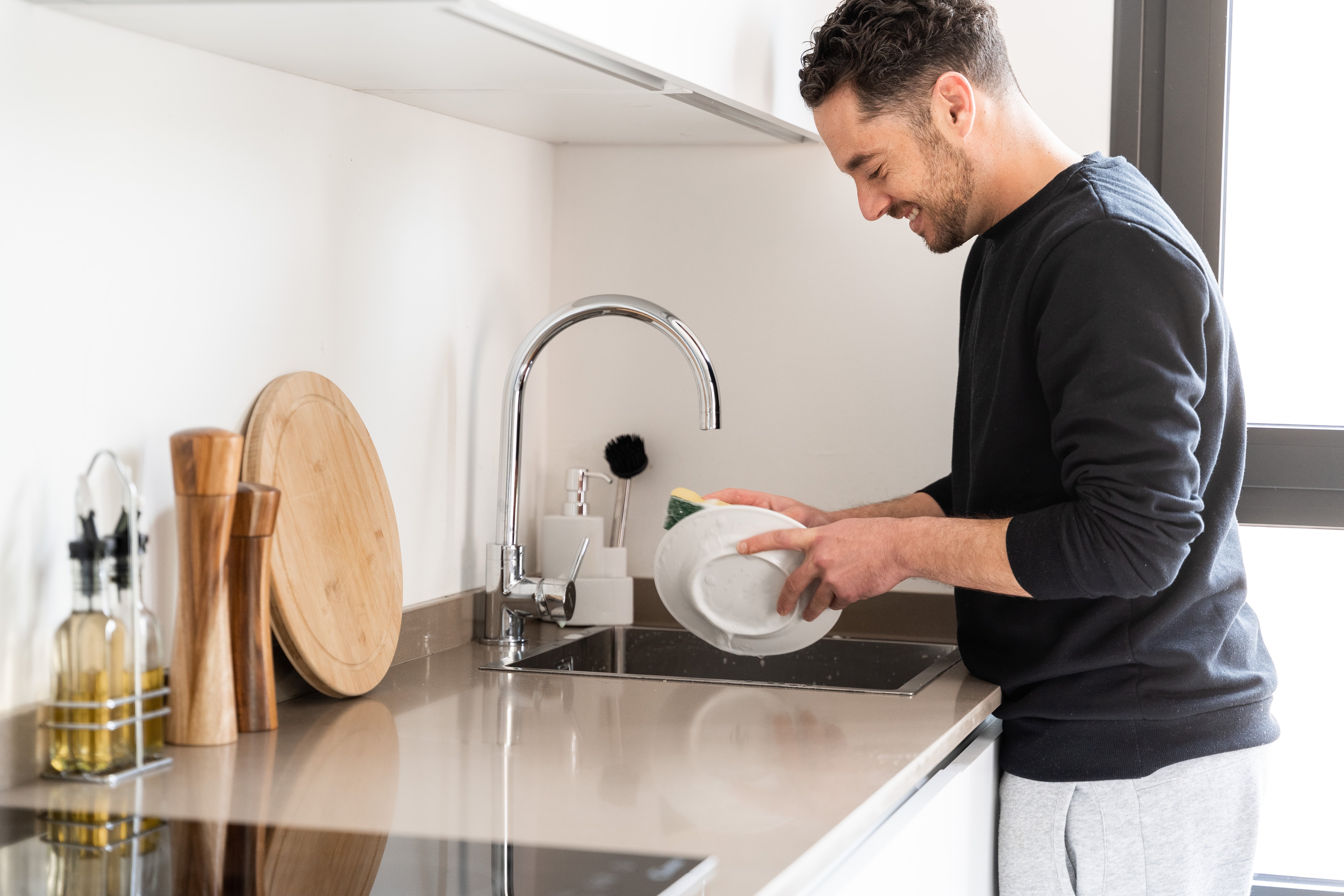 Person smiling while washing dishes at a kitchen sink, wooden cutting board and seasoning bottles are on the counter