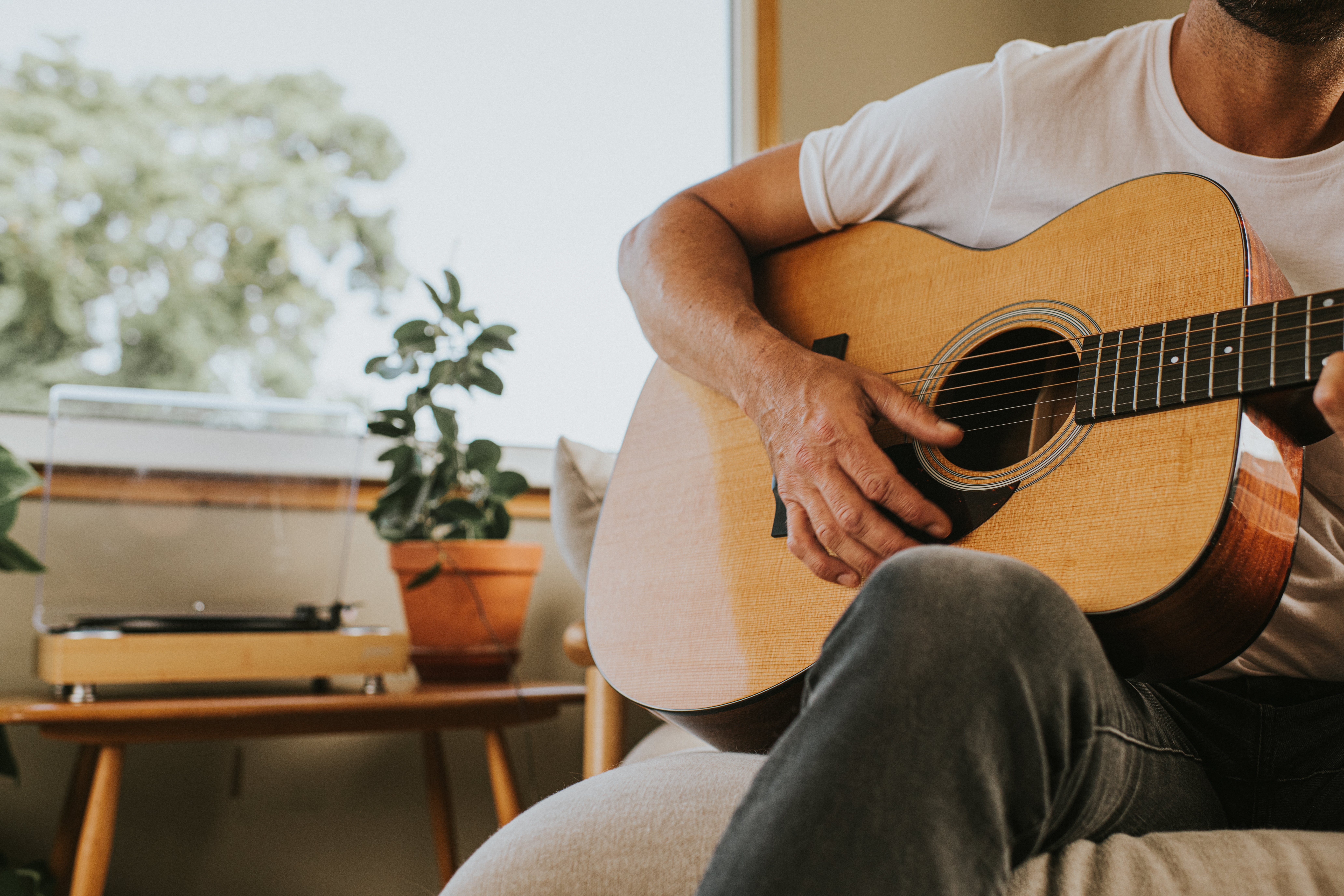 A person strums an acoustic guitar while seated in a cozy room, with a potted plant and a turntable in the background, creating a relaxed atmosphere