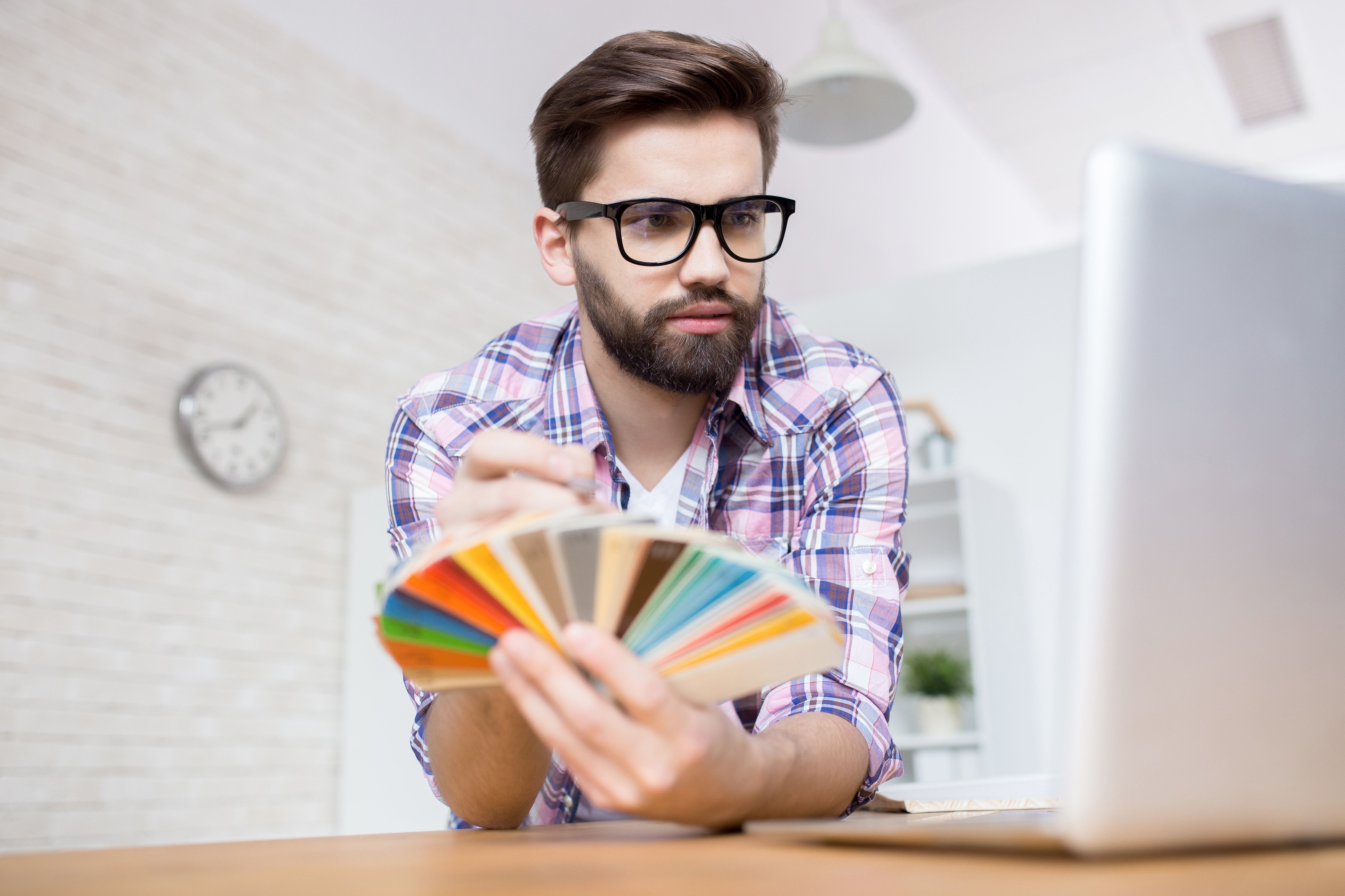 Man with glasses holding a color swatch, looking at a laptop, possibly deciding on paint colors or designs
