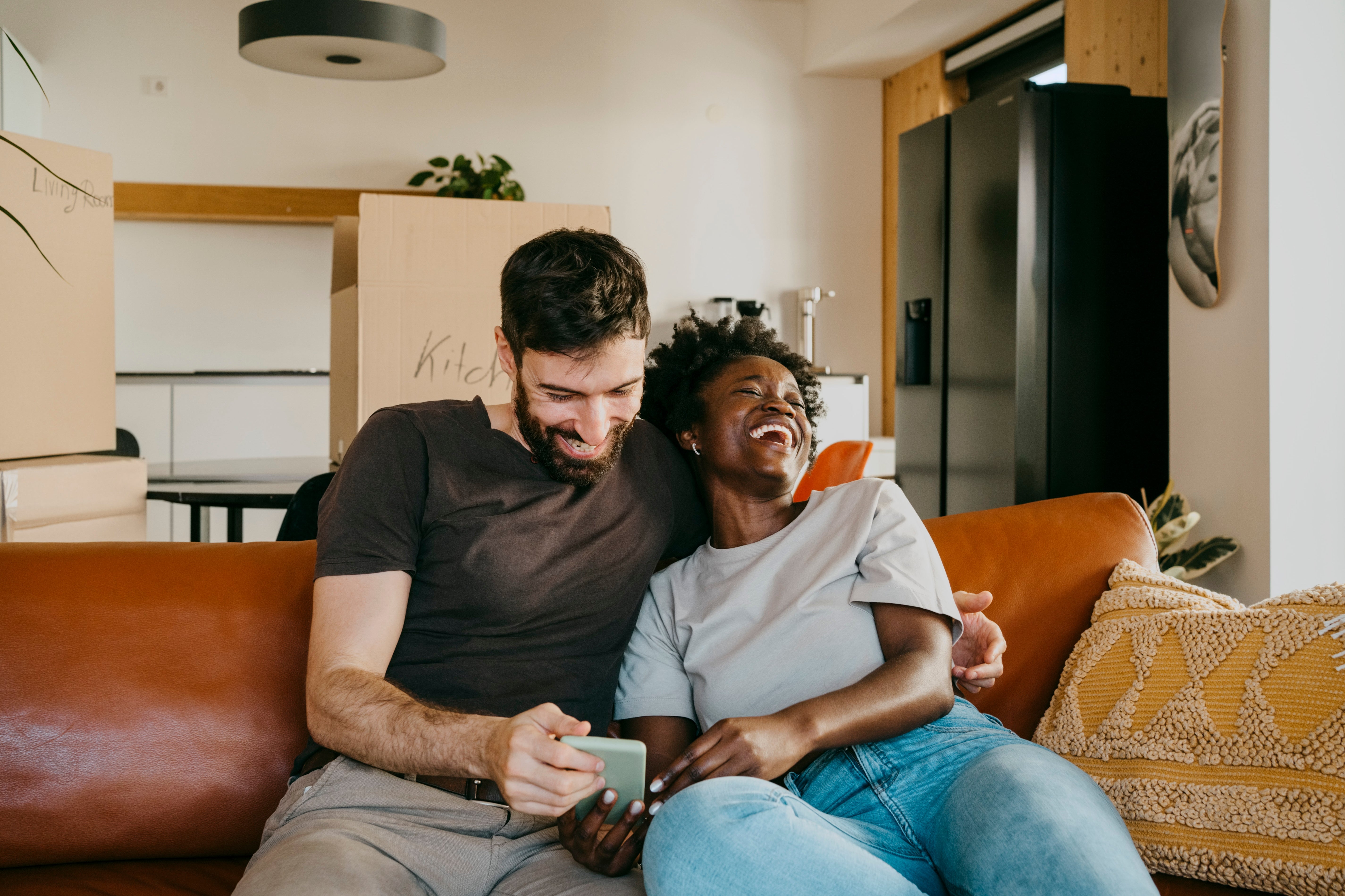 A happy couple sits close on a couch, smiling and looking at a phone together. They appear relaxed and content in a home setting