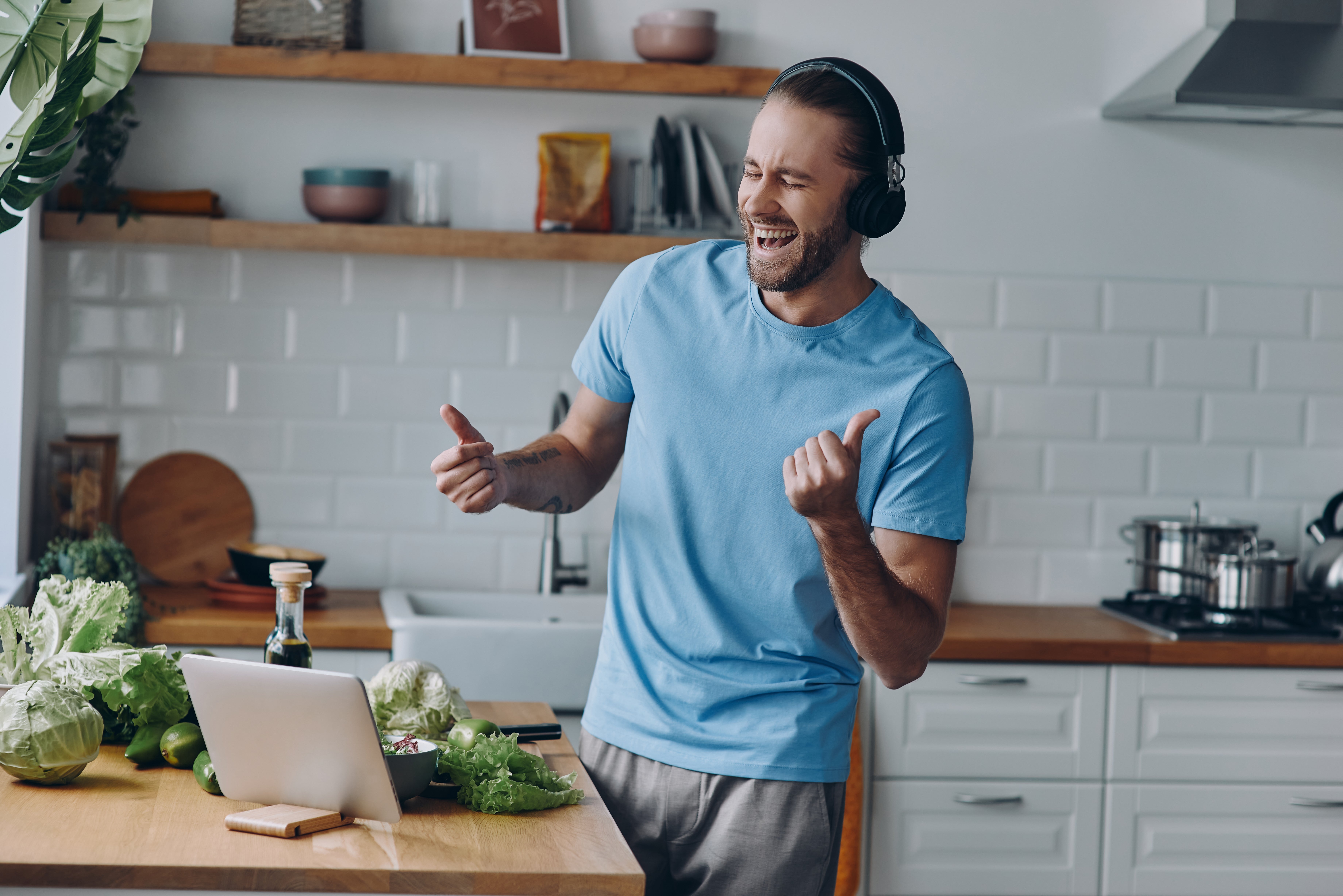 Man wearing headphones, dancing joyfully in a kitchen while looking at a laptop. Fresh vegetables are on the counter