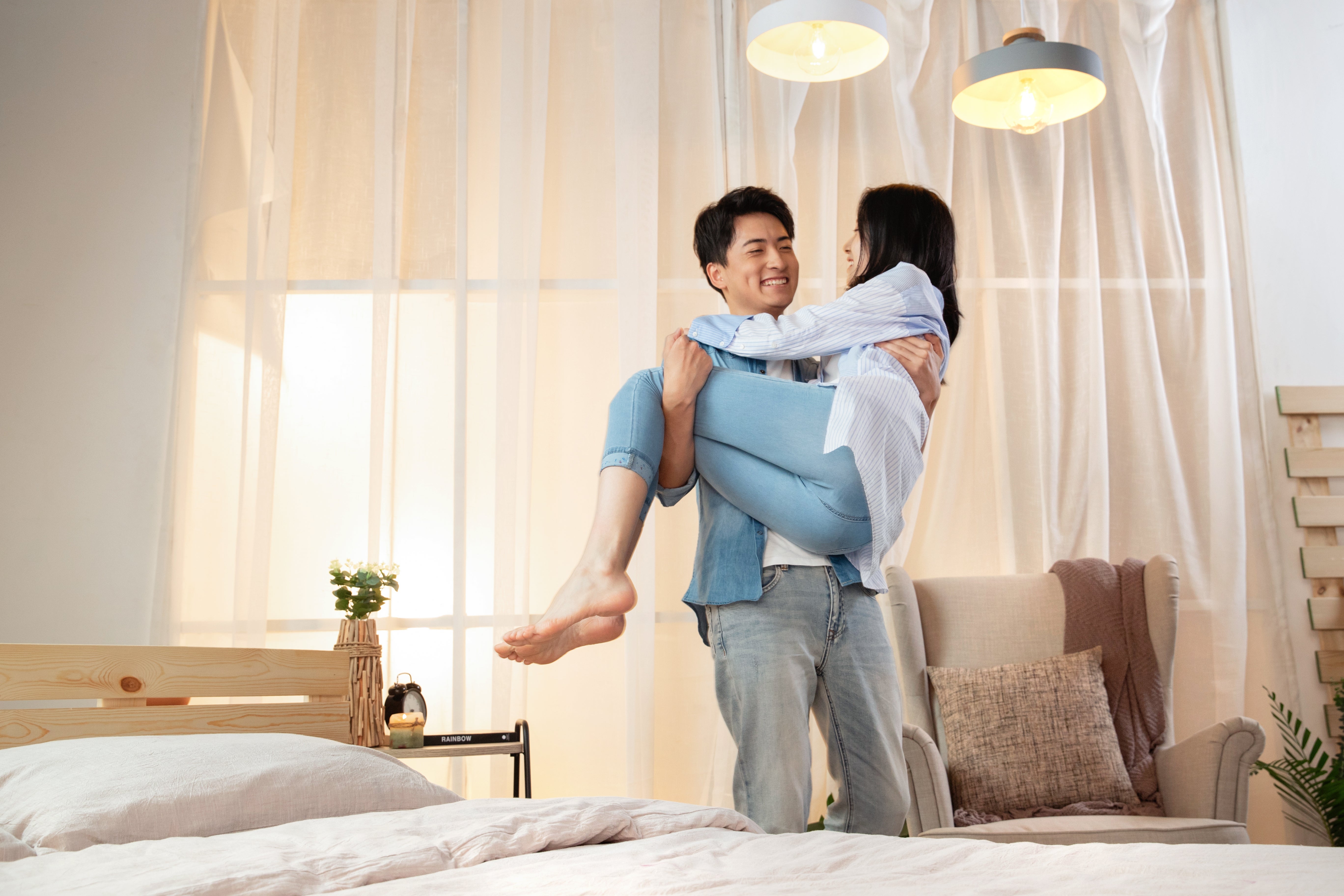 A couple shares a joyful moment as one person lifts the other in a cozy bedroom