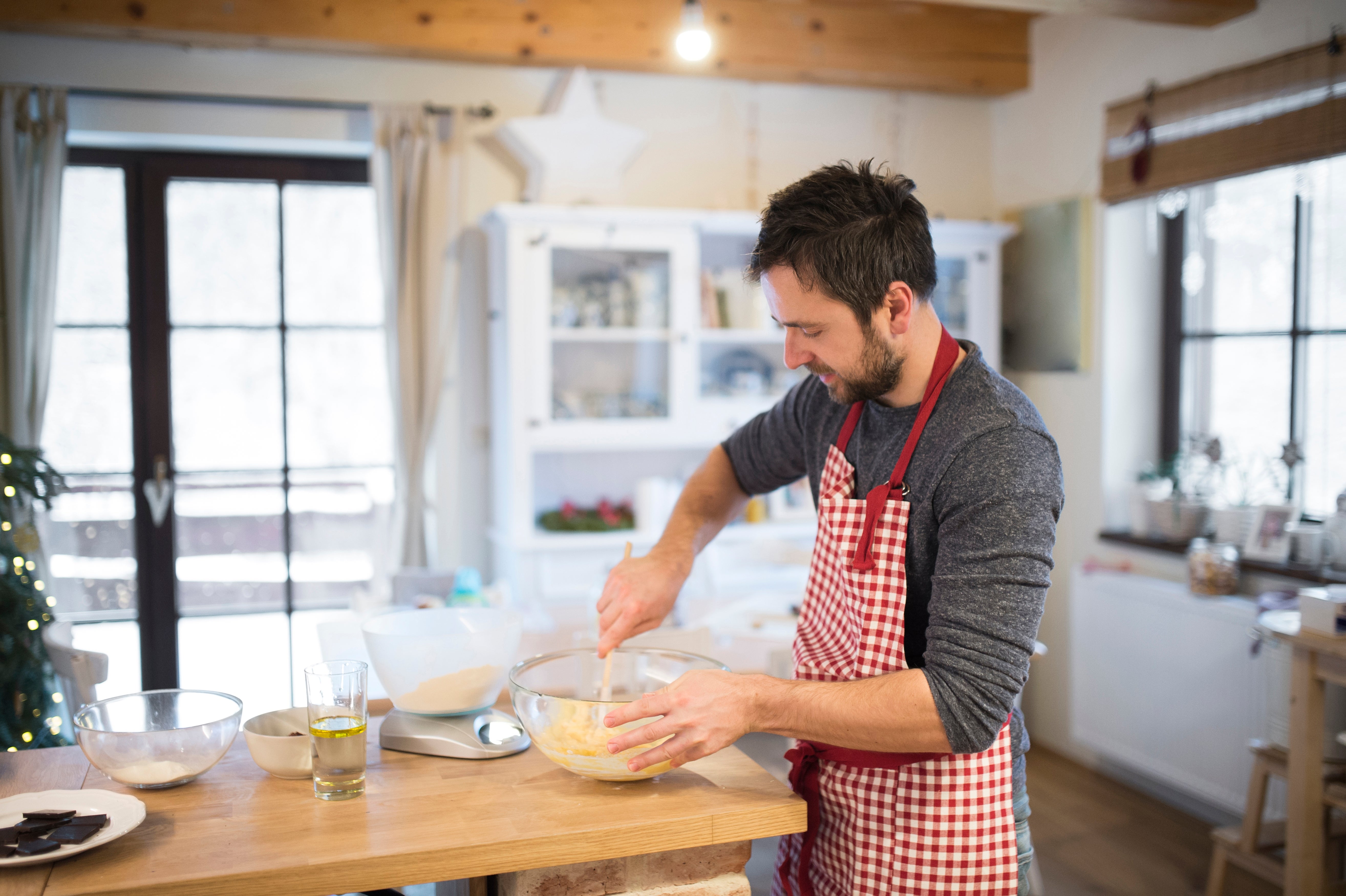 A man in a cozy kitchen, wearing a red checkered apron, mixes ingredients in a bowl, engaging in home baking