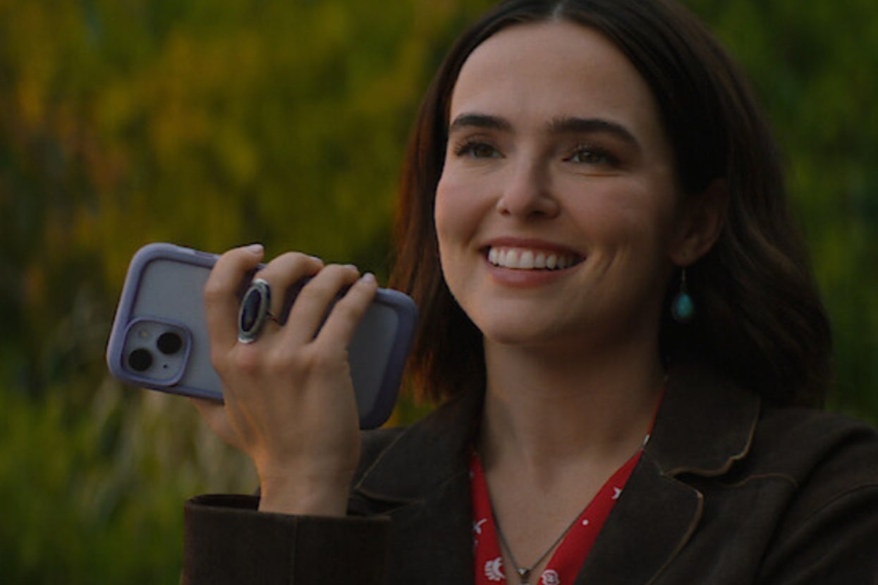 Person smiling, holding a smartphone. They are wearing a dark jacket over a red top